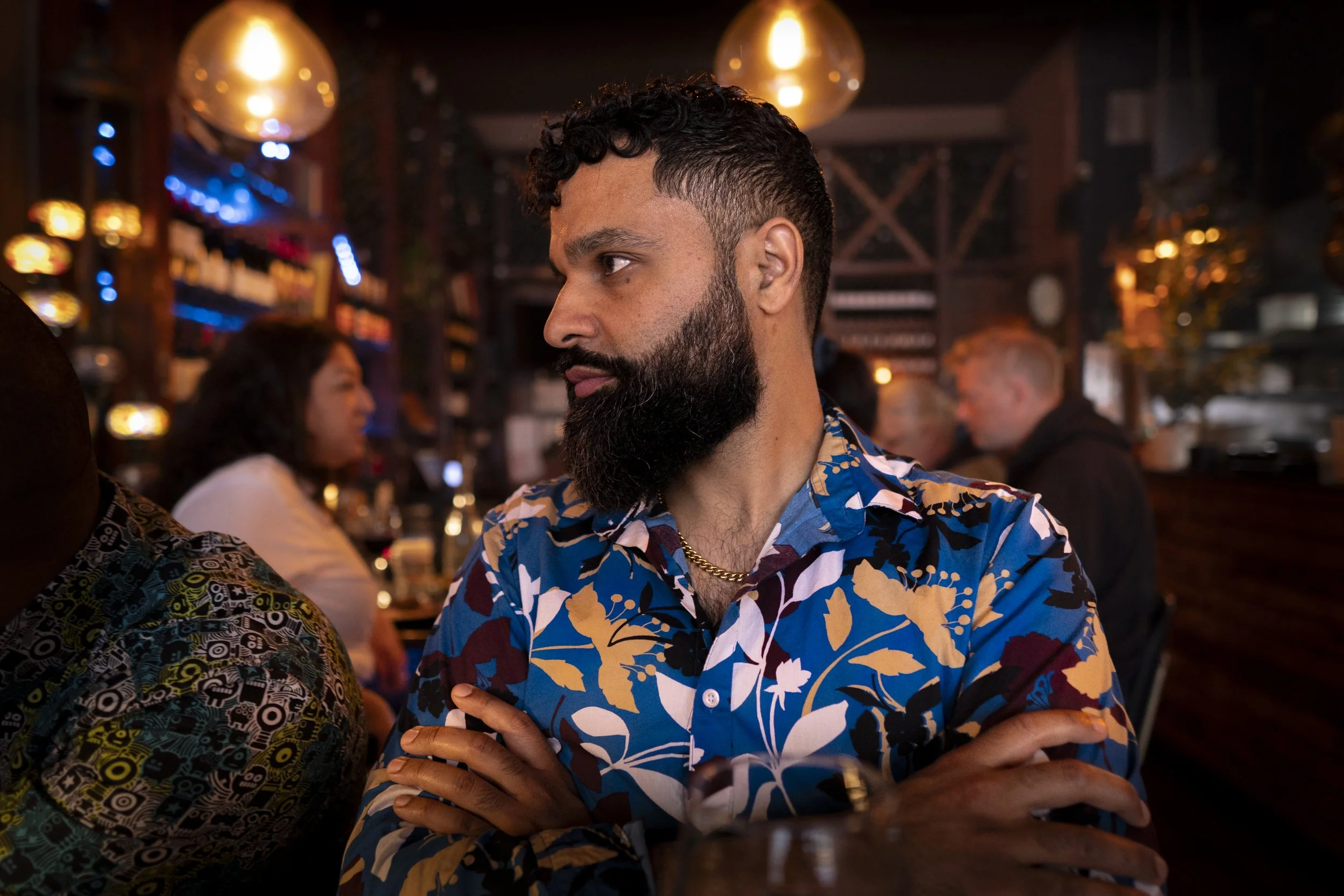 A bearded man with short curly hair wearing a colorful floral shirt and gold chain, standing with arms crossed in a dimly lit bar or restaurant with warm lighting and people in the background.