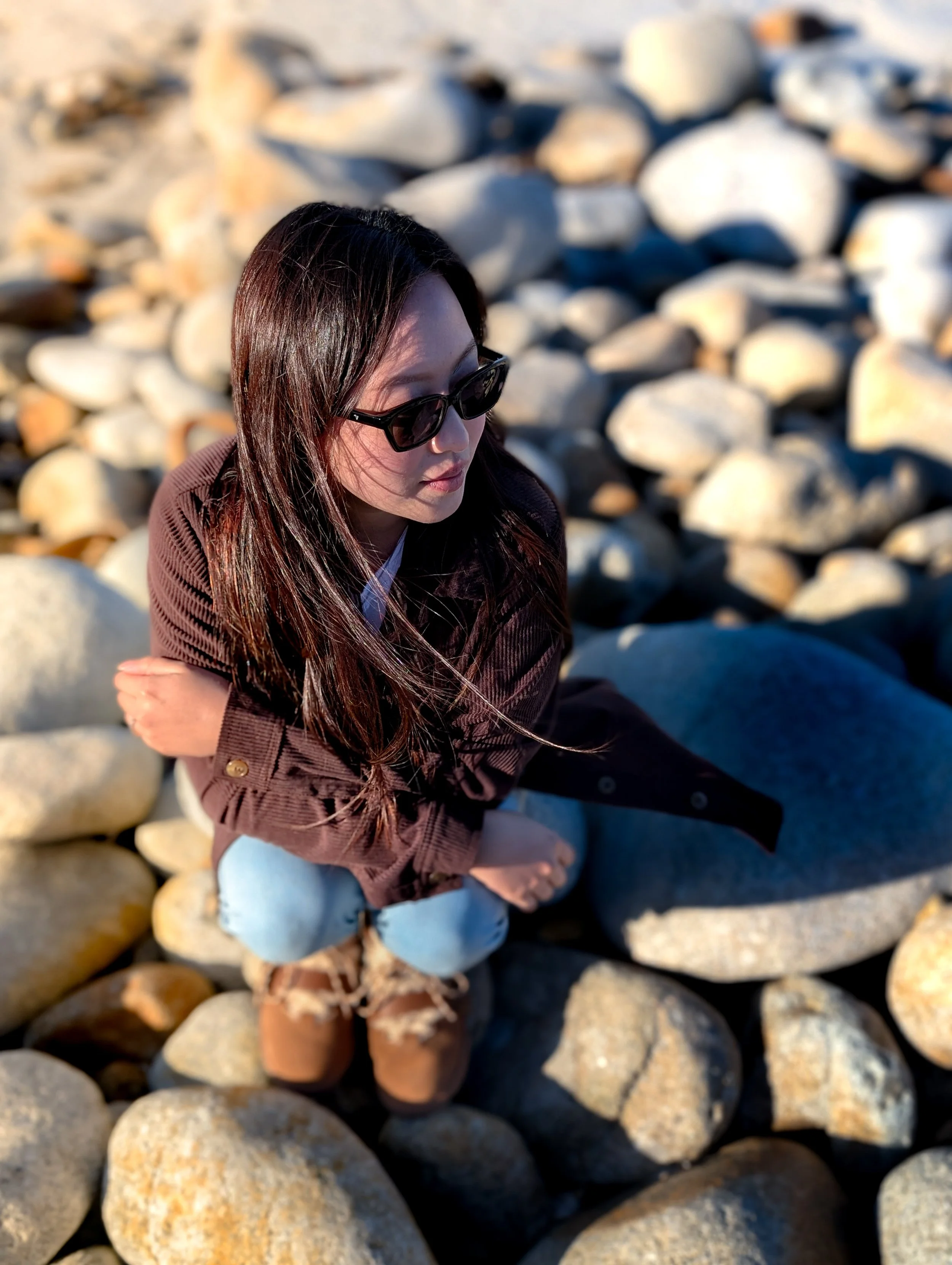A woman with long dark hair, wearing sunglasses, a brown corduroy jacket, and light blue jeans, sitting on a bed of large rocks outside.