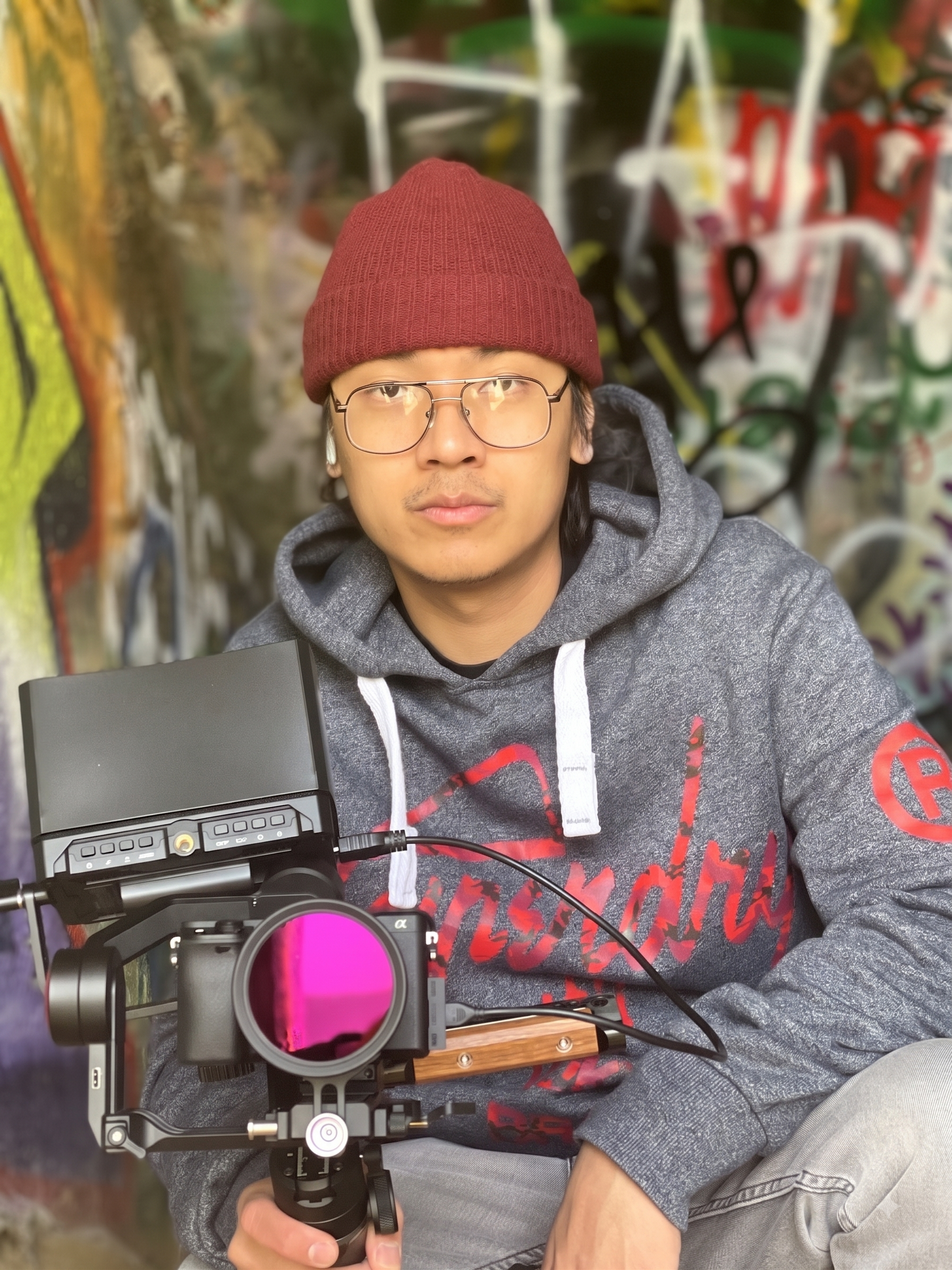 A man wearing glasses, a red beanie, and a gray hoodie, operating a camera stabilizer in front of a graffiti-covered wall.