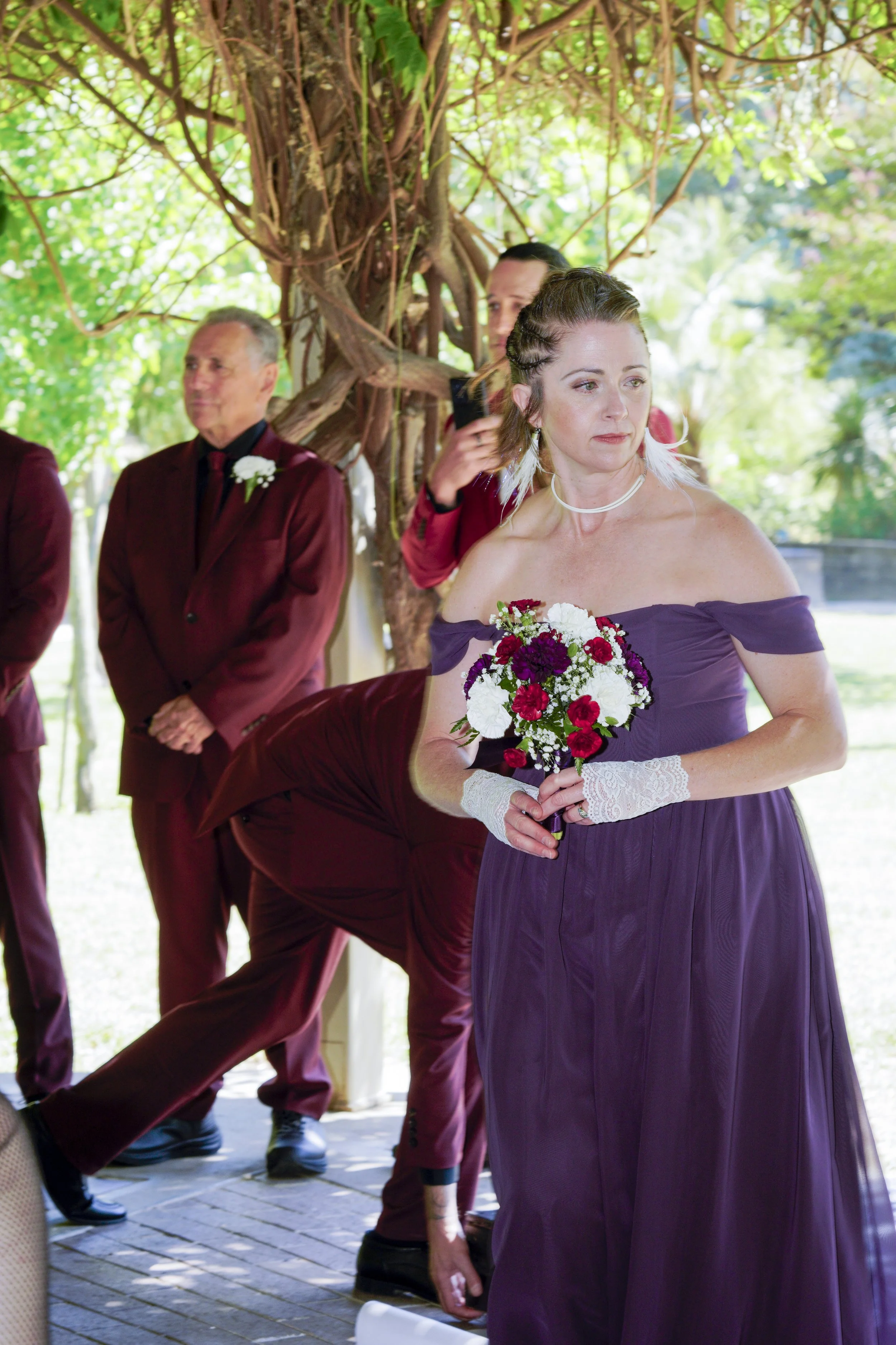 A woman in a purple dress holding a bouquet of red, white, and purple flowers, standing outdoors under a tree, with people dressed in burgundy suits behind her.