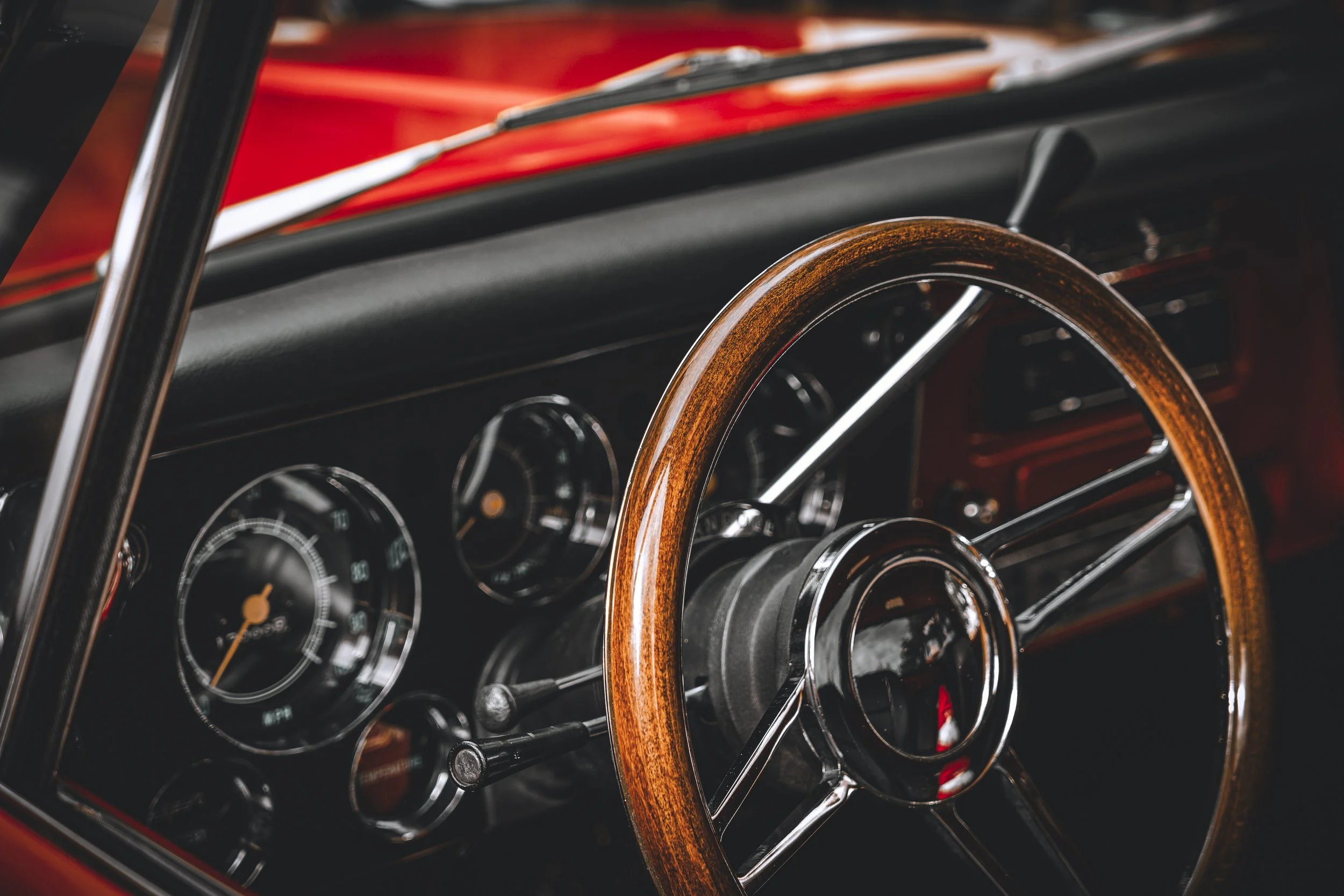 Close-up of a vintage car interior showing a brown wooden steering wheel, black dashboard with analog gauges, and a red body exterior.