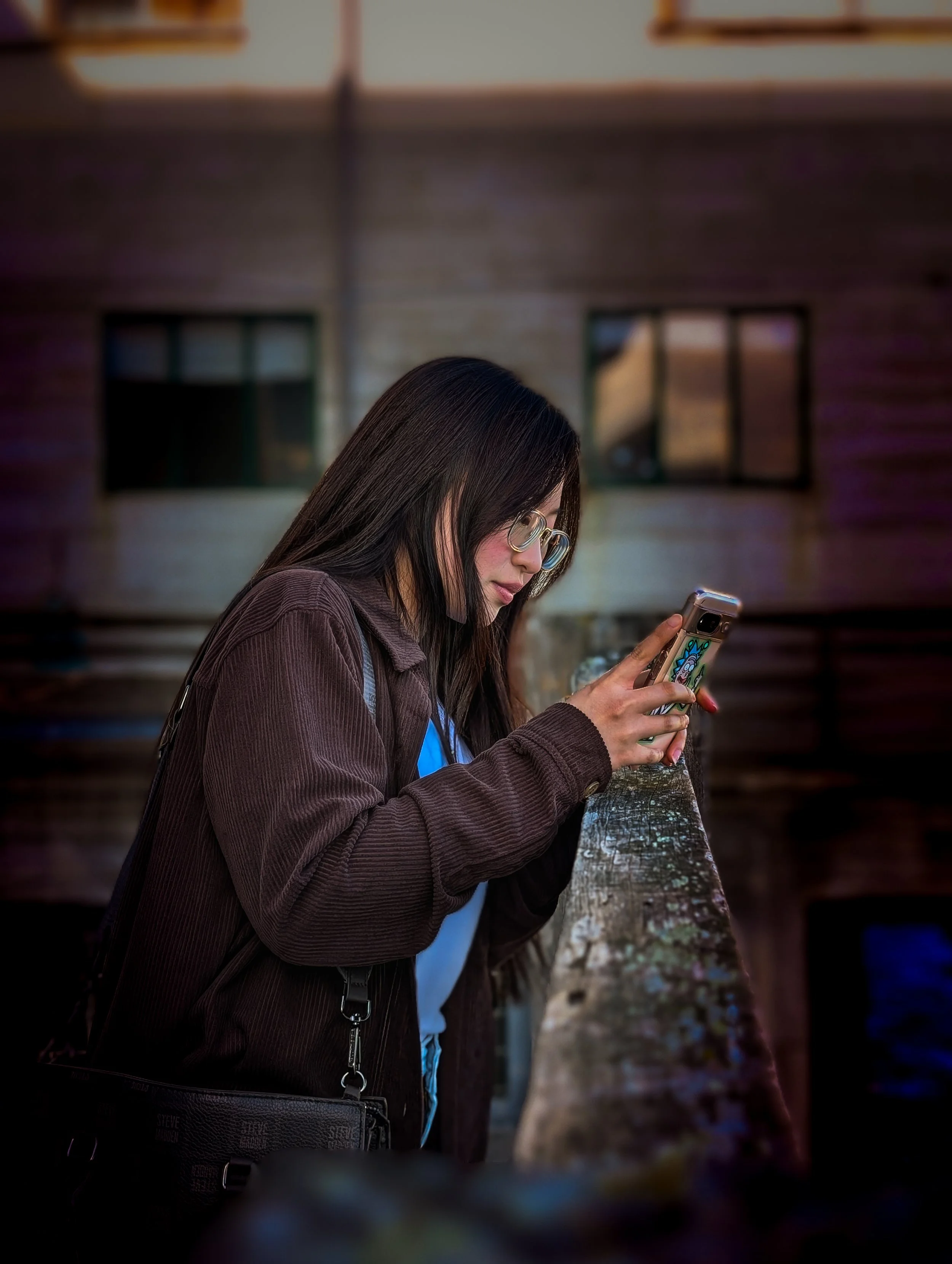 A woman with long dark hair, glasses, and a blue shirt, wearing a brown jacket, is looking at her phone while leaning on a wooden railing outdoors, with a blurred house in the background.