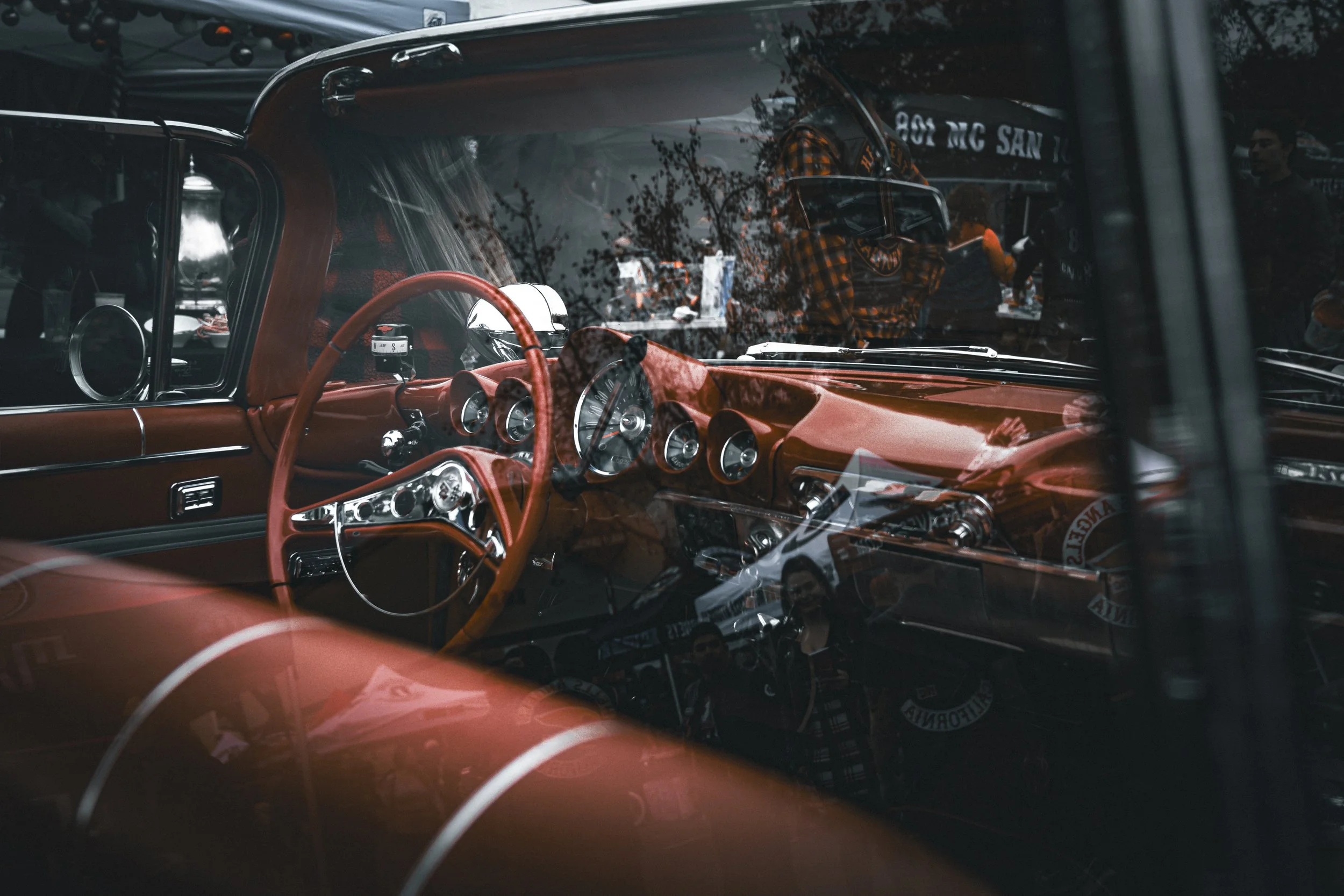 Photo of the interior of a vintage red car, showing the dashboard, steering wheel, and gauges, with reflections of trees and people on the windshield.