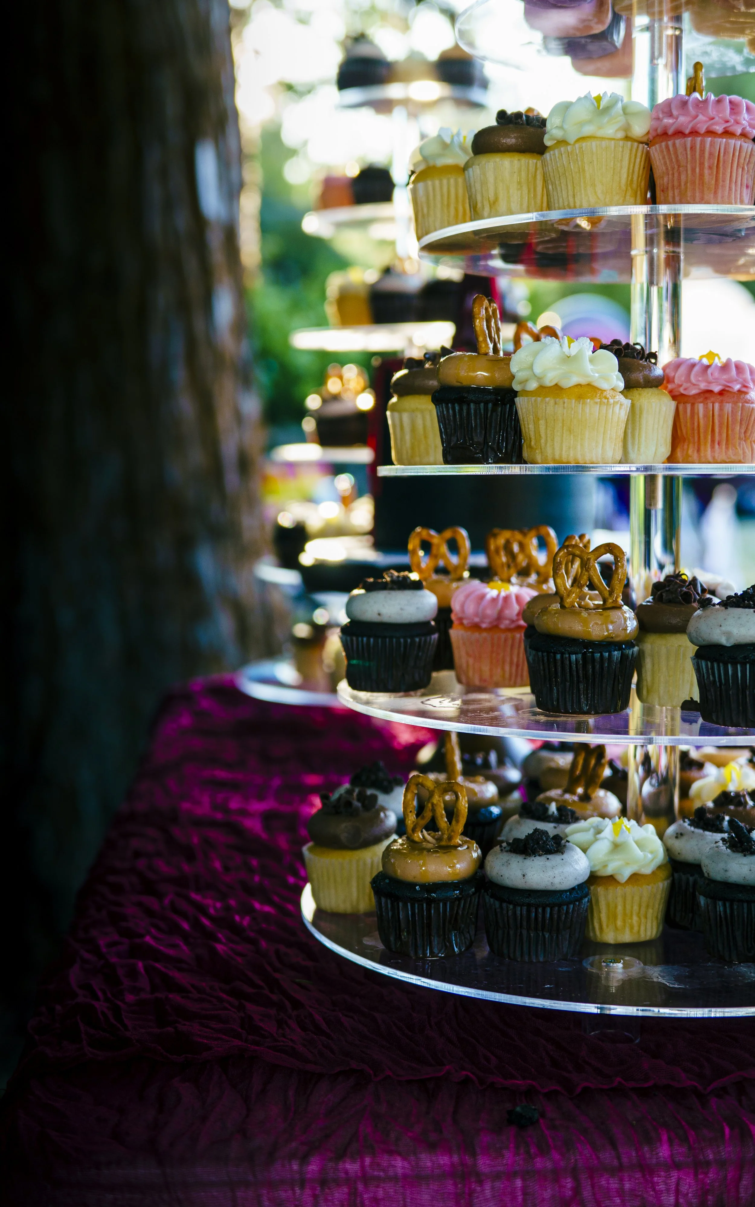 A display of colorful cupcakes on a tiered stand at an outdoor event, with a blurred background of trees and bright sunlight.
