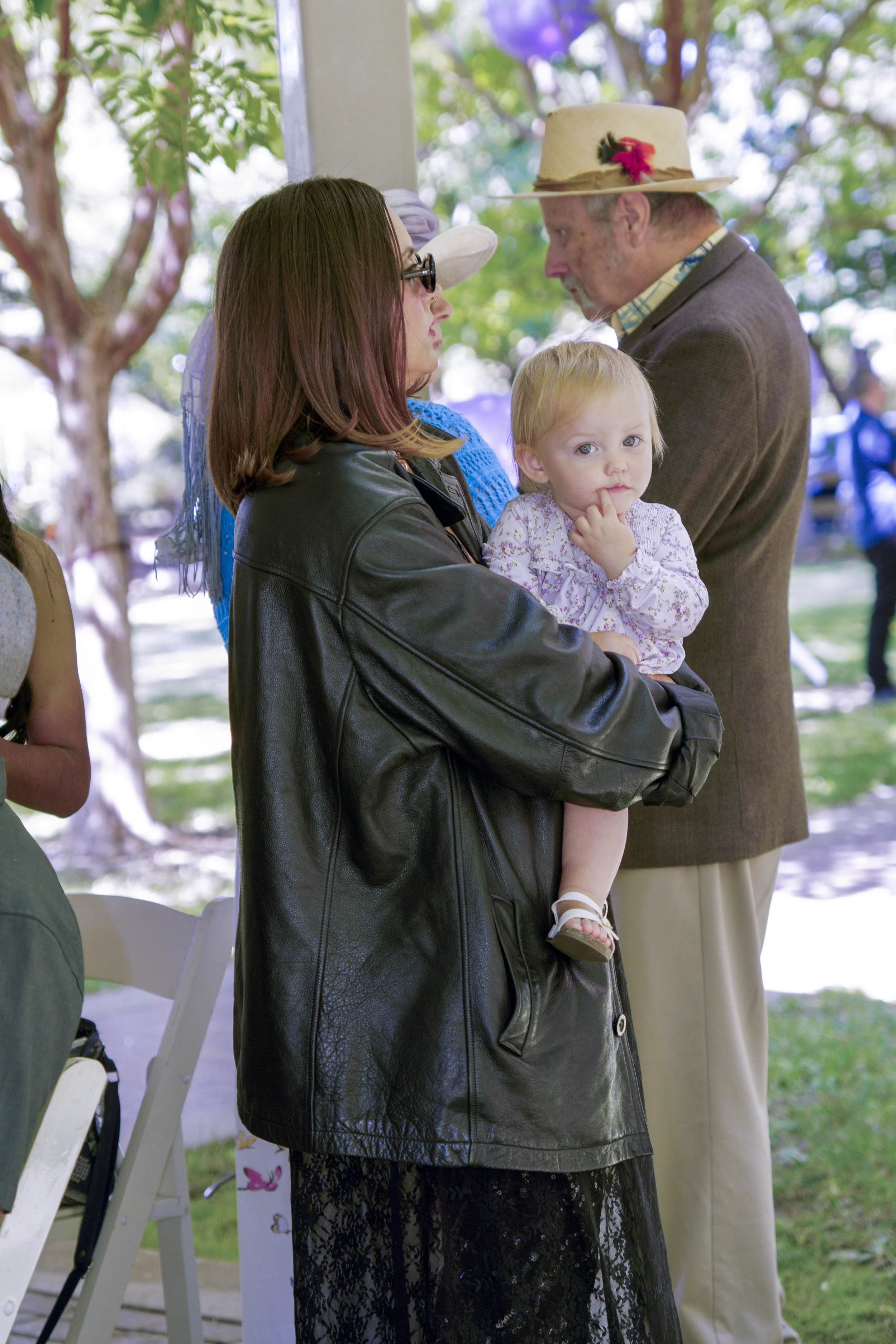 Woman holding a young girl outdoors, facing an elderly man wearing a straw hat with pink flower and black ribbon, in a park with trees.