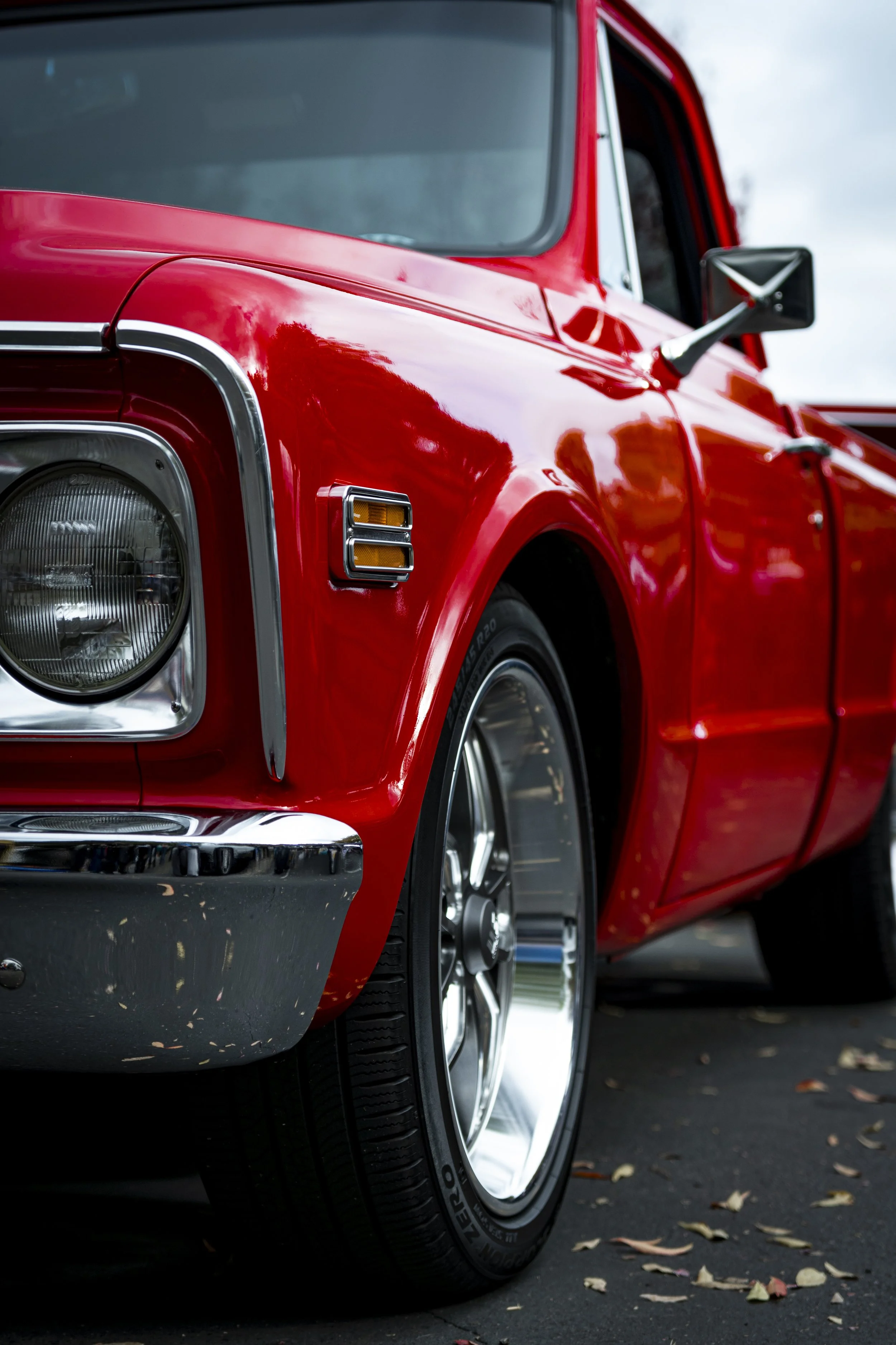 Close-up of the front part of a vintage red car, showing the headlight, wheel, and side mirror, parked on an asphalt surface with scattered fallen leaves.