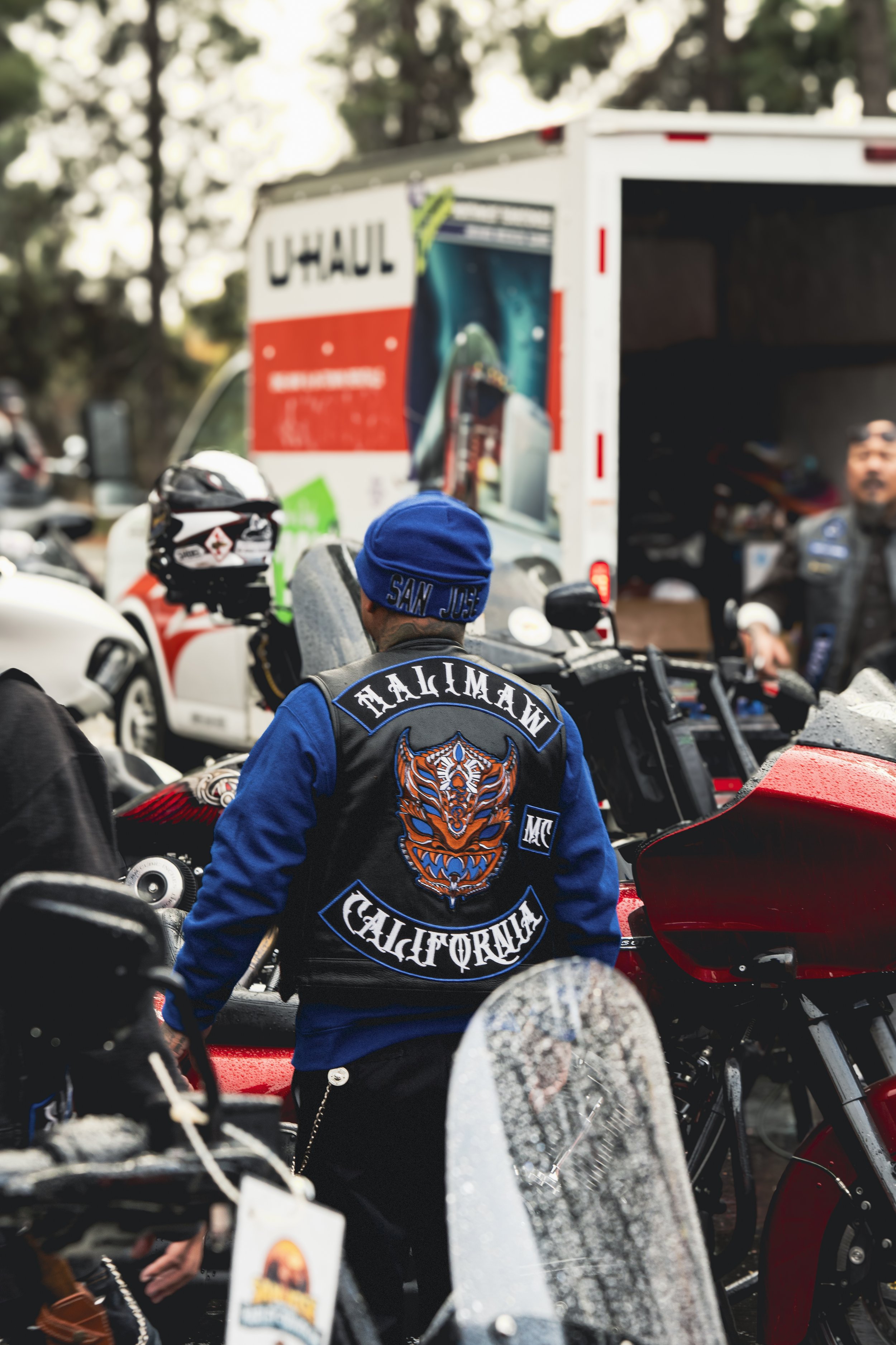 A man wearing a California motorcycle club jacket and a blue beanie standing among motorcycles outside, with a U-Haul truck in the background.