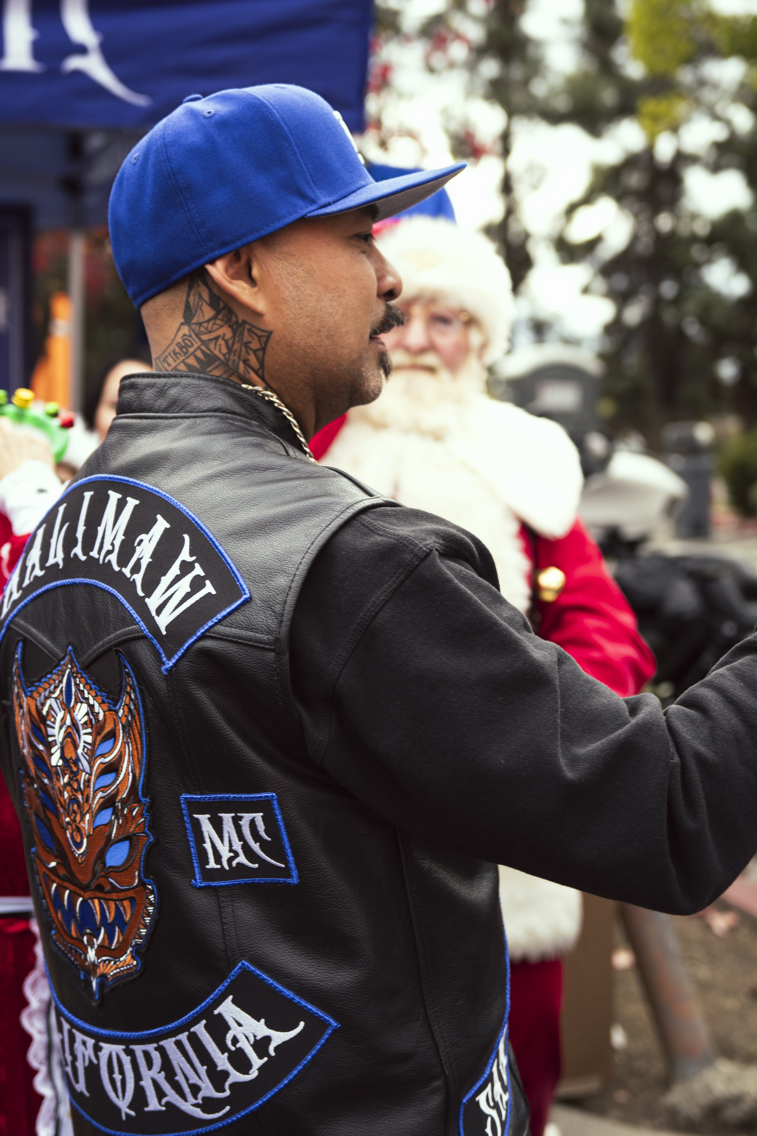 A man wearing a black leather jacket with colorful embroidered patches and a blue baseball cap, standing outdoors during a festive event with Christmas decorations and a person dressed as Santa Claus in the background.