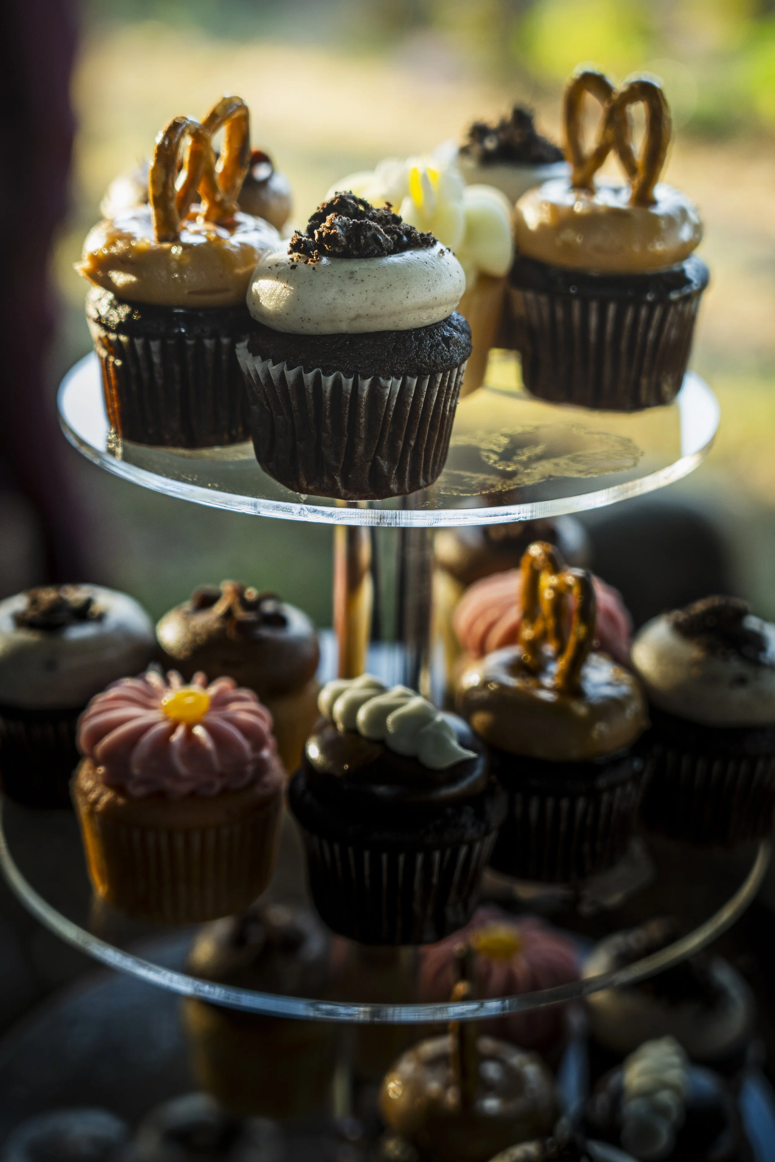 Assorted decorated cupcakes on a glass stand, with various toppings and decorations, in an outdoor setting with blurred background.