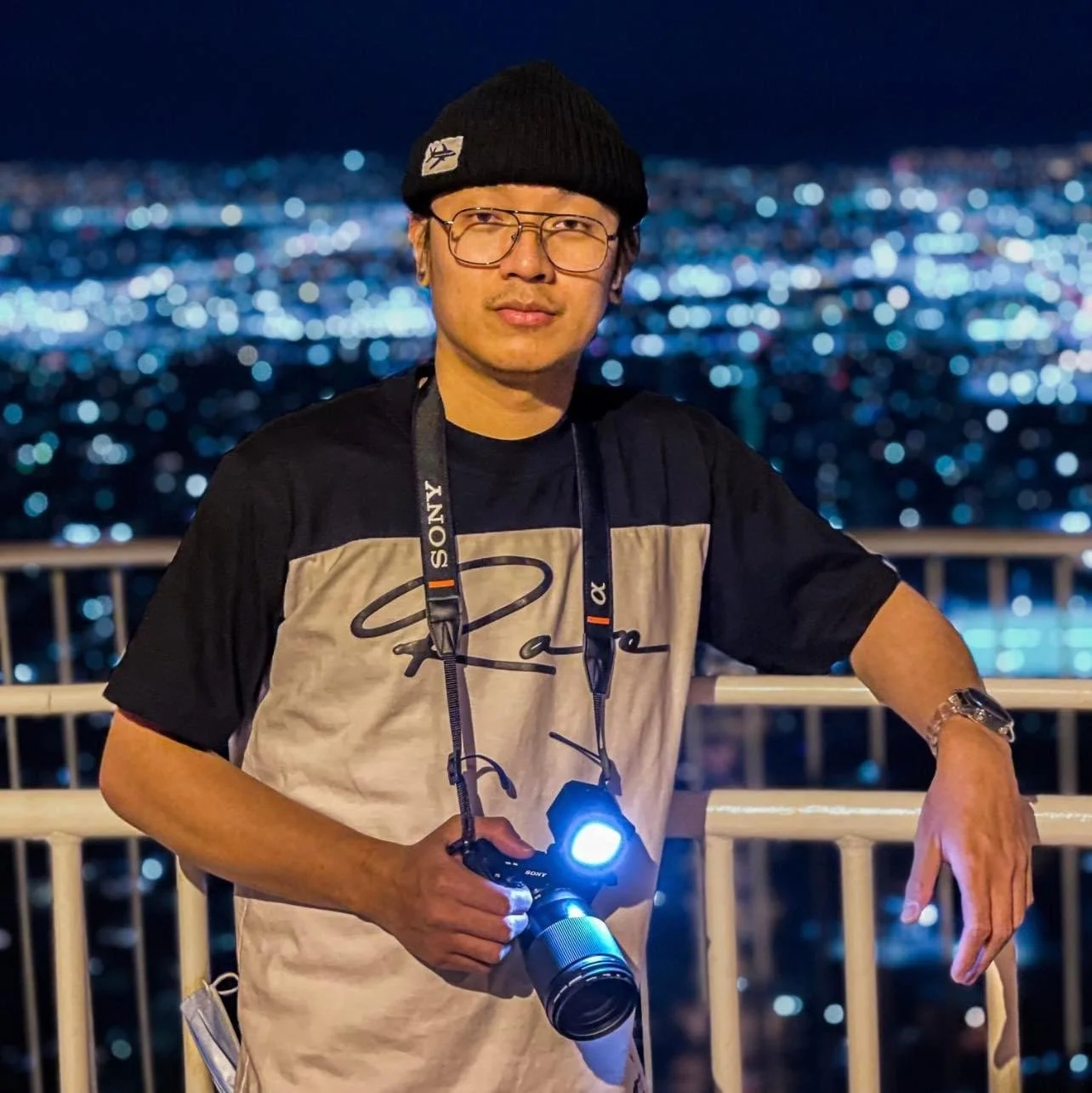 A man wearing glasses, a black beanie, and a signature T-shirt holding a camera with a flashlight, standing against a cityscape at night.