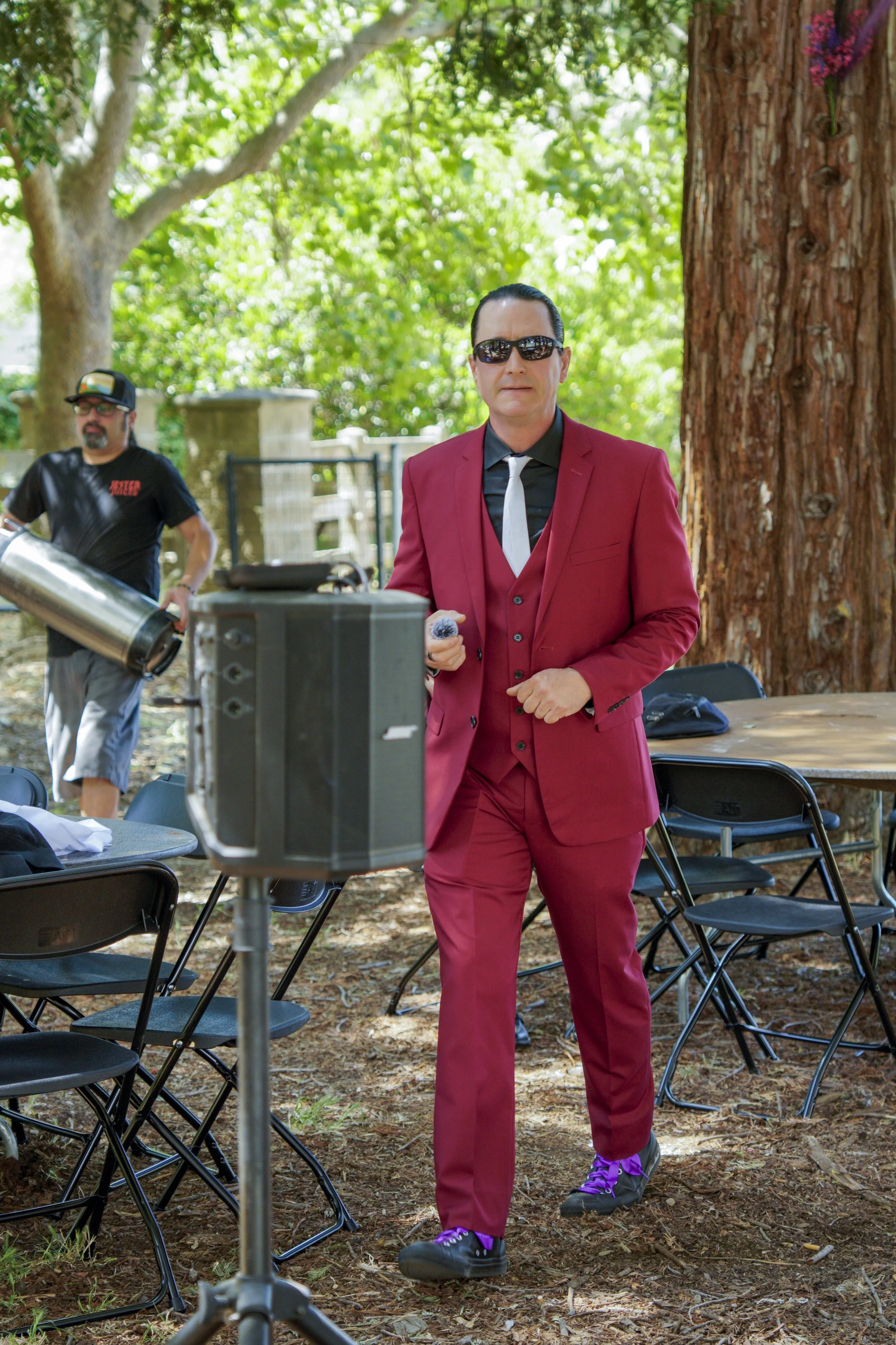 A man in a red suit, black shirt, black tie, sunglasses, and purple sneakers standing outdoors under trees with green leaves, near a large tree trunk, with screening equipment and chairs around him.