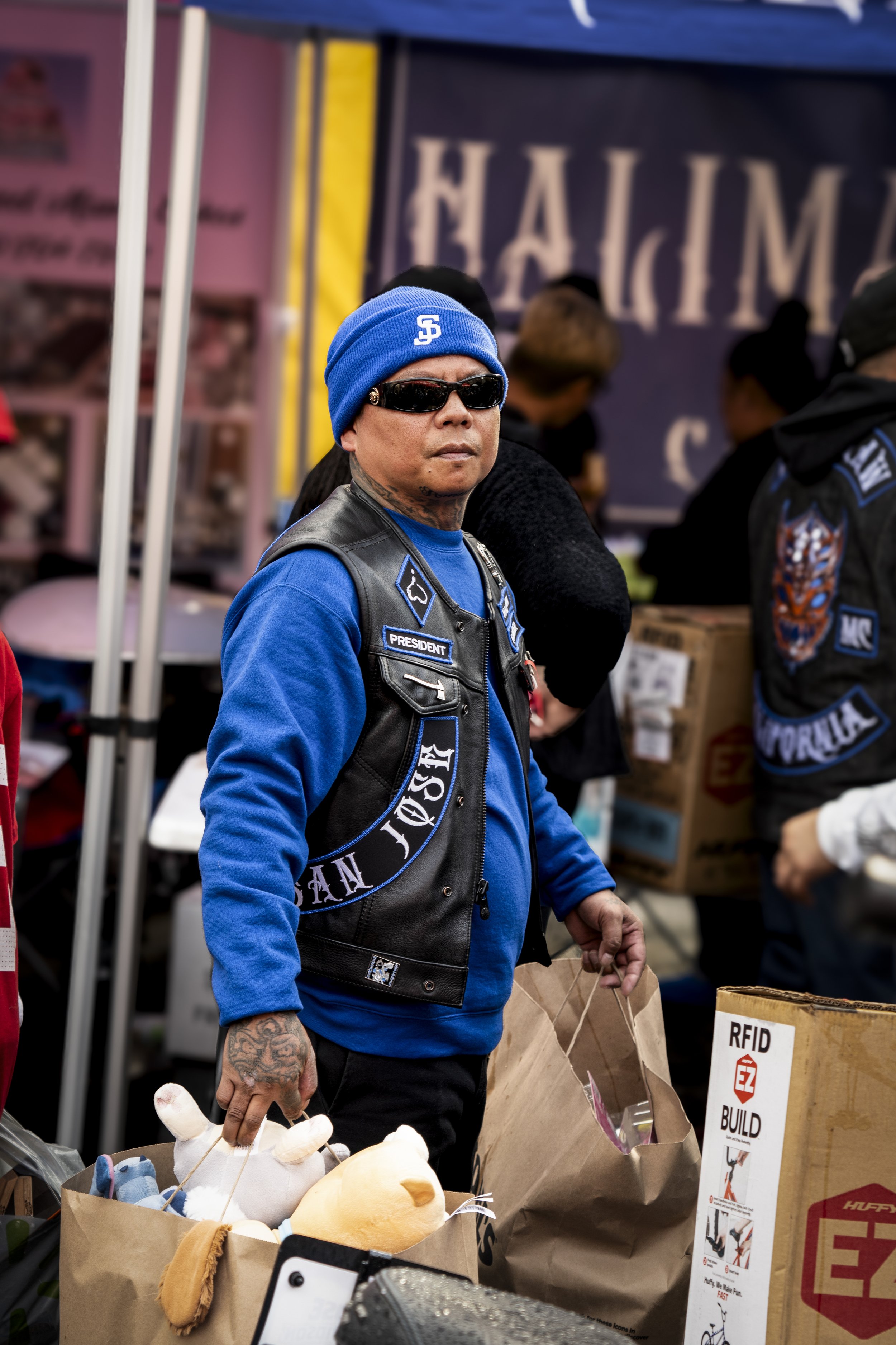 A man with tattoos, wearing sunglasses, a blue beanie, and a black leather vest with patches, is at an outdoor market. He is holding a shopping bag and there are stuffed animals on a table in front of him.
