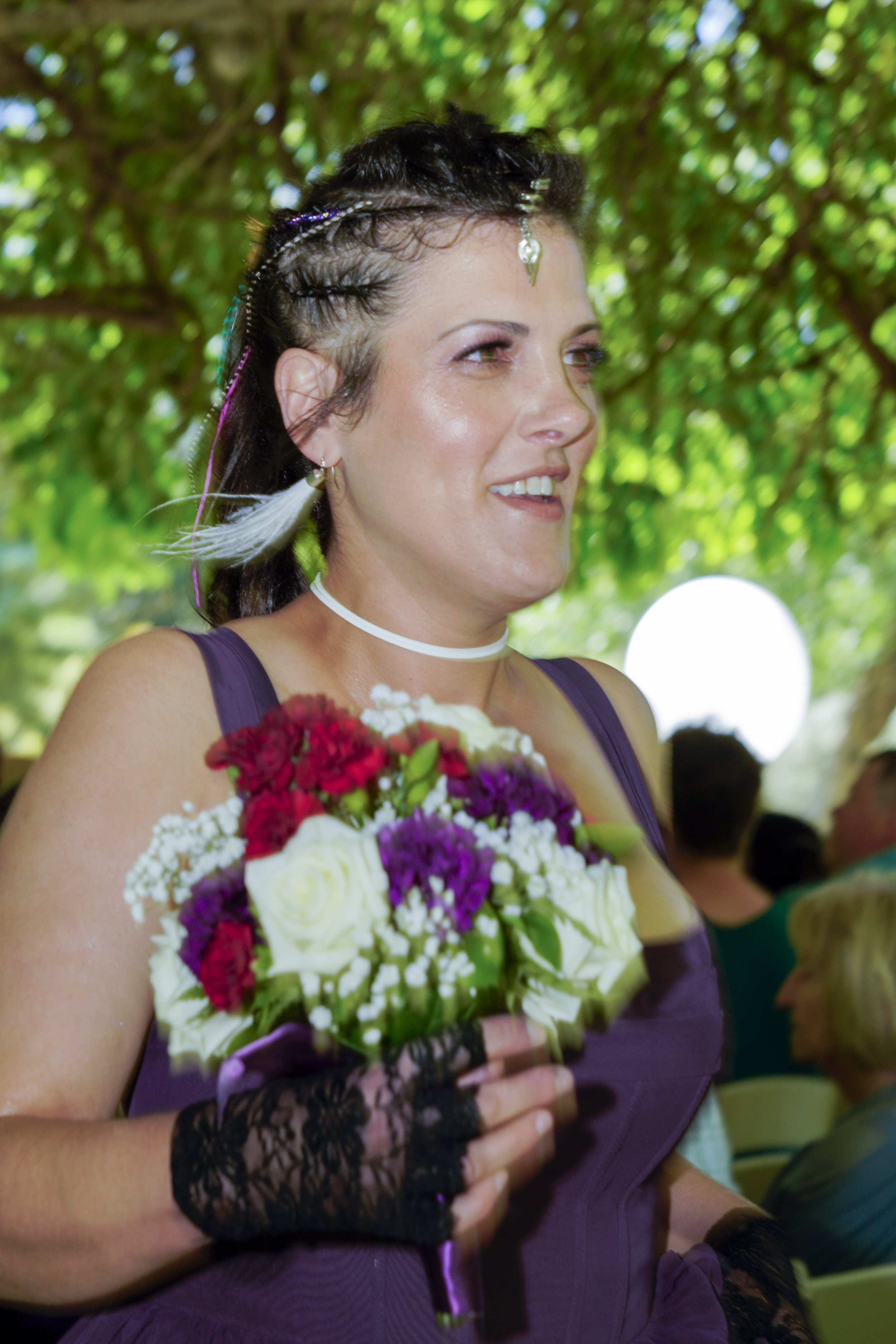 A woman with braided hair adorned with beads and a jewel, holding a bouquet of colorful flowers, wearing a purple dress and black lace gloves, at an outdoor event.