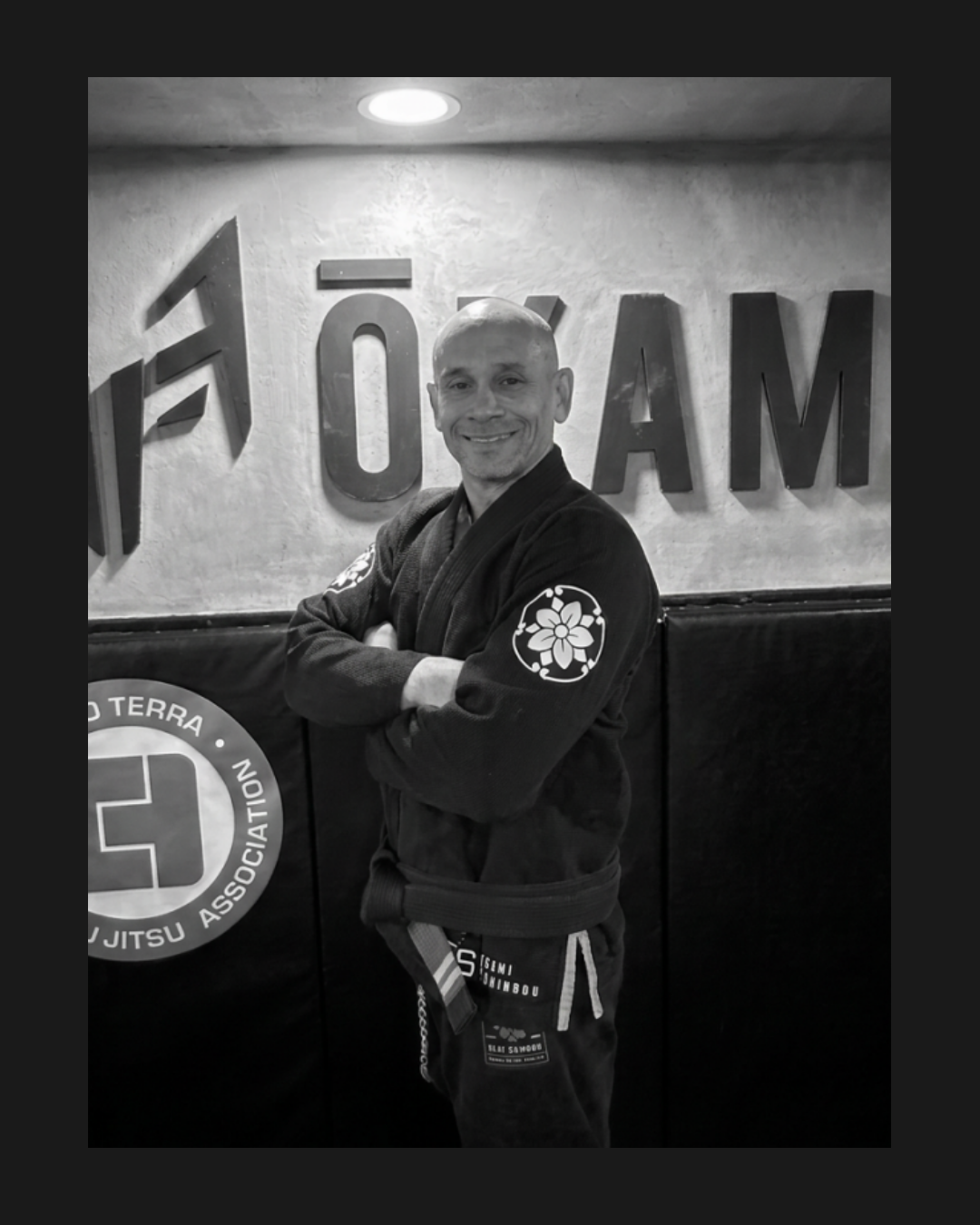 Hombre en uniforme de jiu jitsu con fondo de pared con letras y logotipo, sonriendo con brazos cruzados, en un gimnasio de artes marciales.