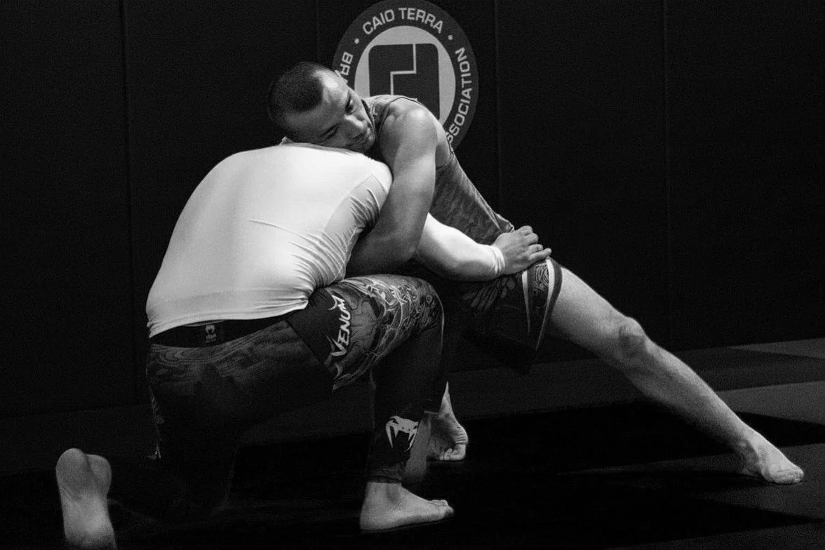 Dos personas practicando jiu-jitsu en un gimnasio, en una posición de lucha en el tapete, con un fondo que muestra un logotipo del Club Caio Terra.