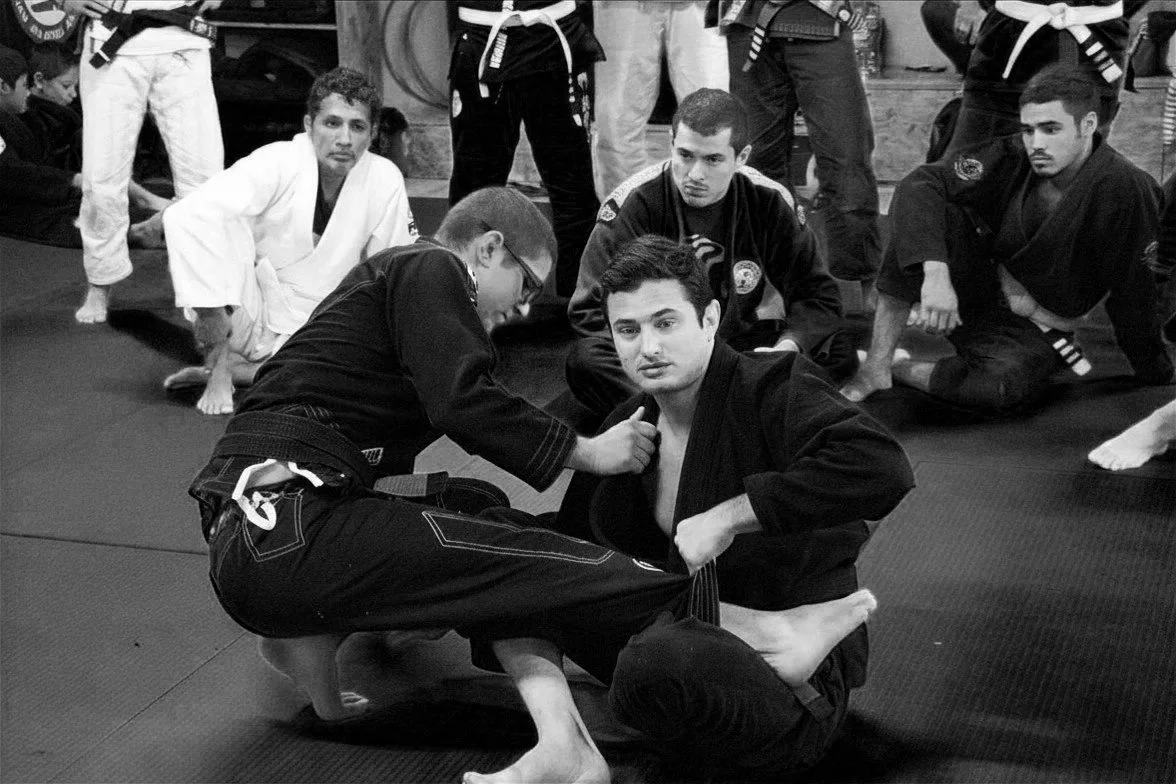 Hombres practicando jiu-jitsu en un gimnasio, con uno al centro en posición de guardia mientras otro le ajusta la parte superior de su kimono.