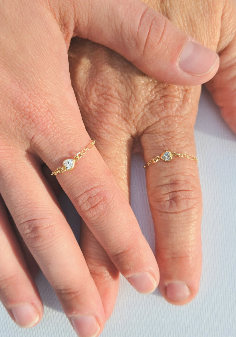 Two hands with gold rings featuring heart-shaped designs and small gemstones, resting on a light surface.
