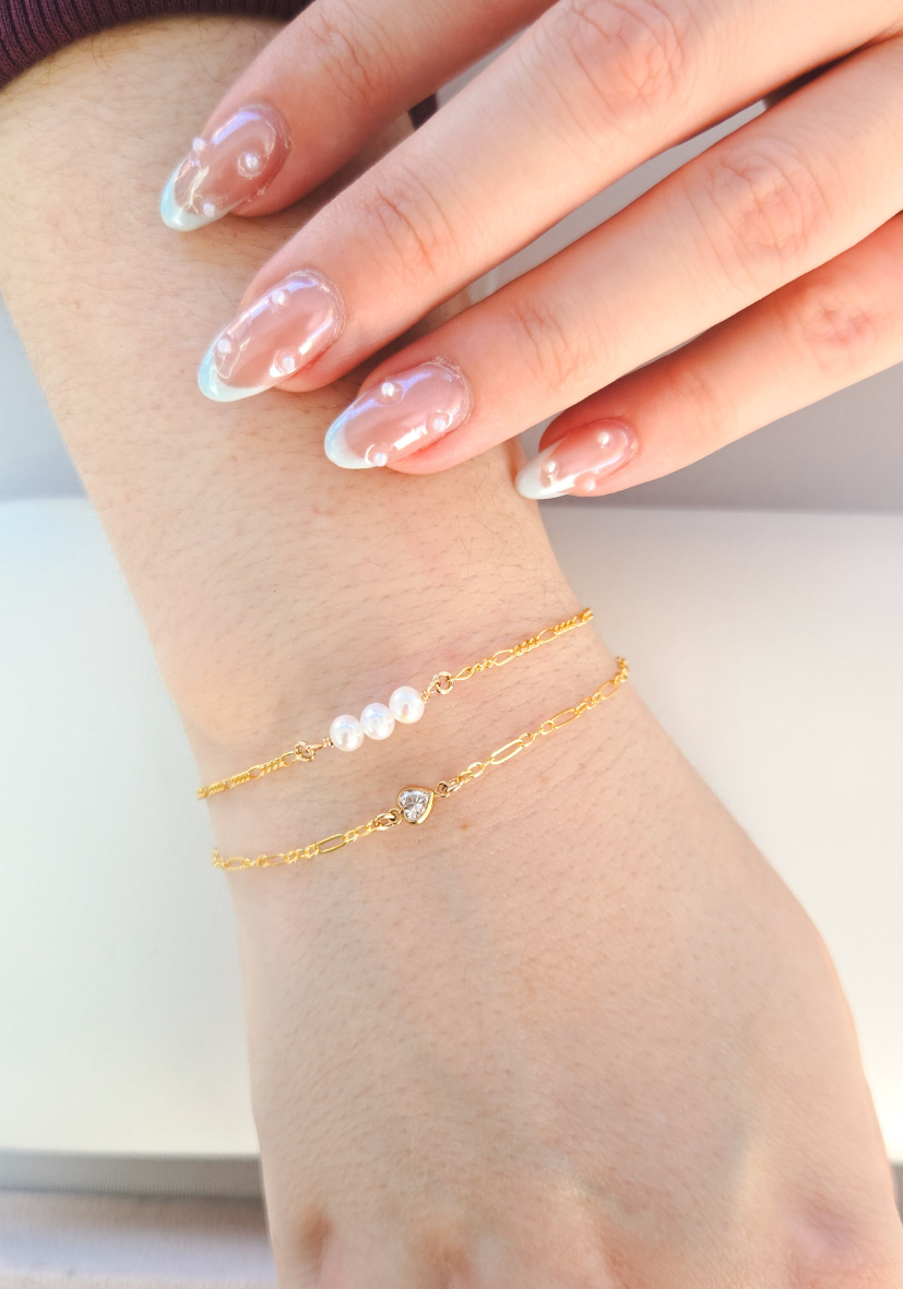 Close-up of a woman's wrist with two gold bracelets, one with pearls and a heart-shaped charm, and the other with a chain link design, with her hand resting on her arm.