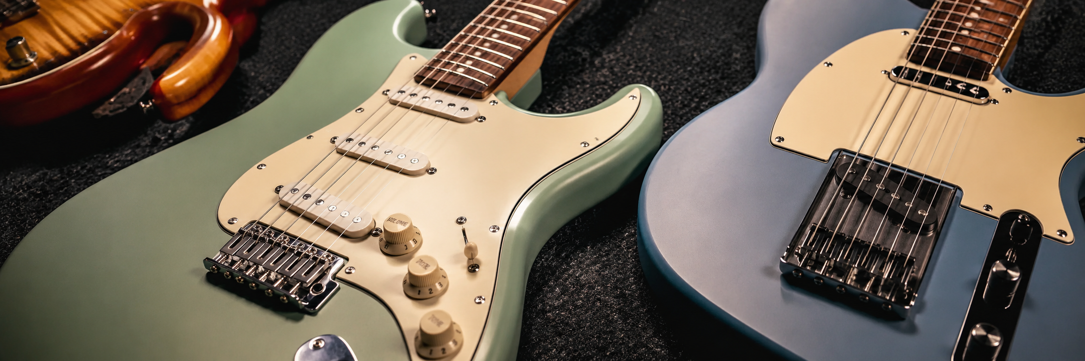 Two electric guitars, one green and one blue, lying side by side on a dark surface, with parts of other guitars visible in the background.