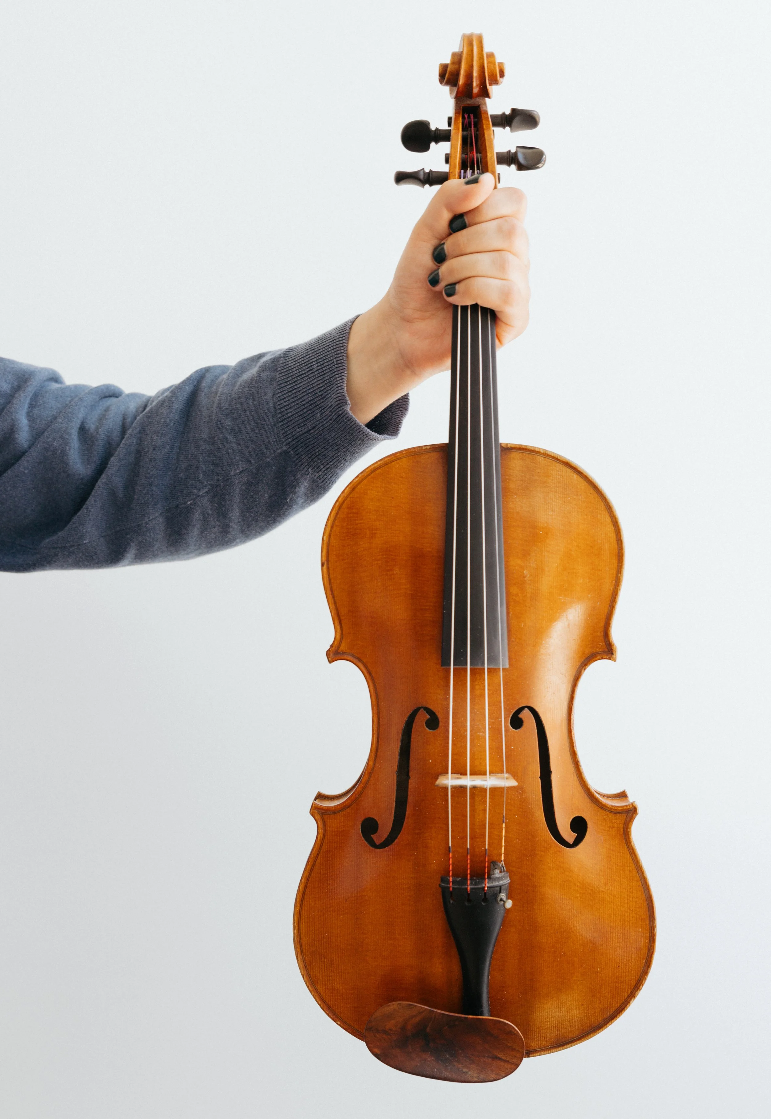 A person wearing a gray long-sleeve shirt holding a wooden violin against a plain white background.