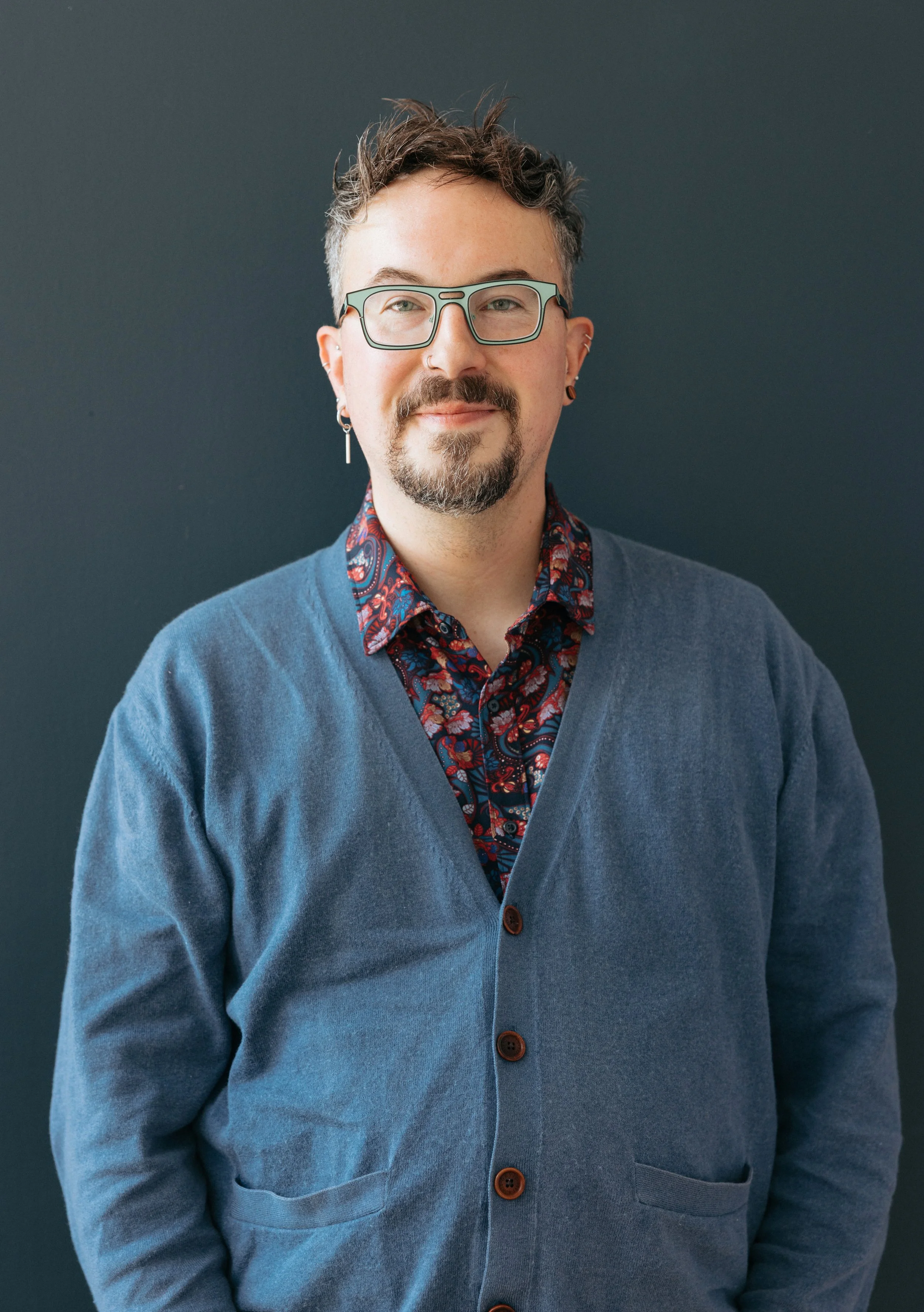 A man with glasses, multiple earrings, and a beard wearing a patterned shirt under a blue cardigan against a dark background.