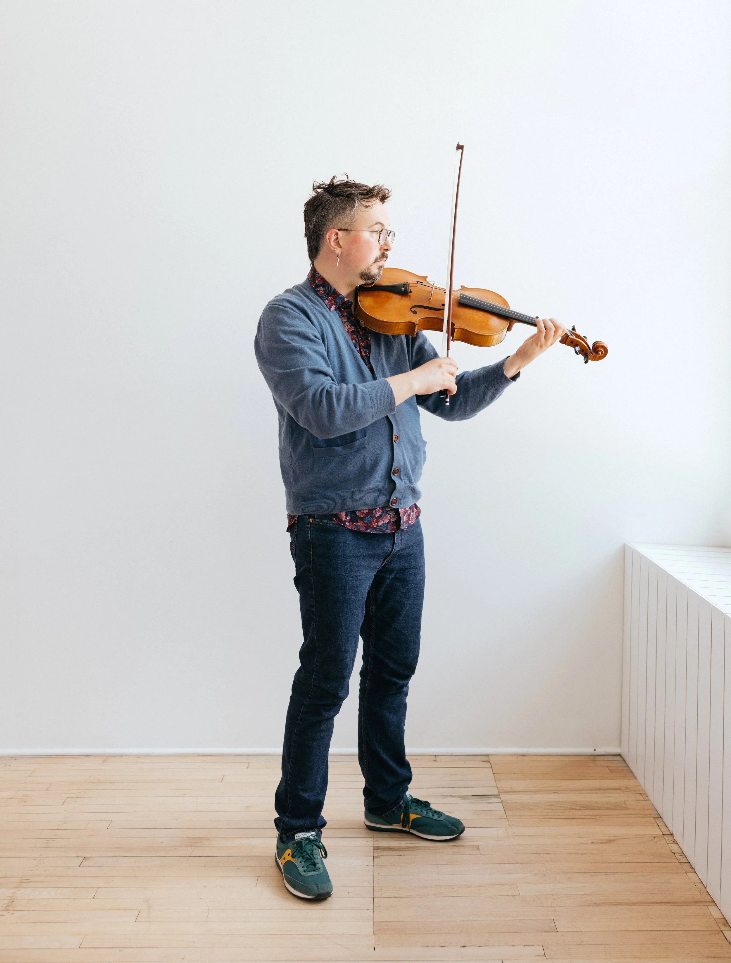 A man playing a violin indoors against a plain white wall, wearing glasses, a blue cardigan, dark jeans, and teal and yellow sneakers.