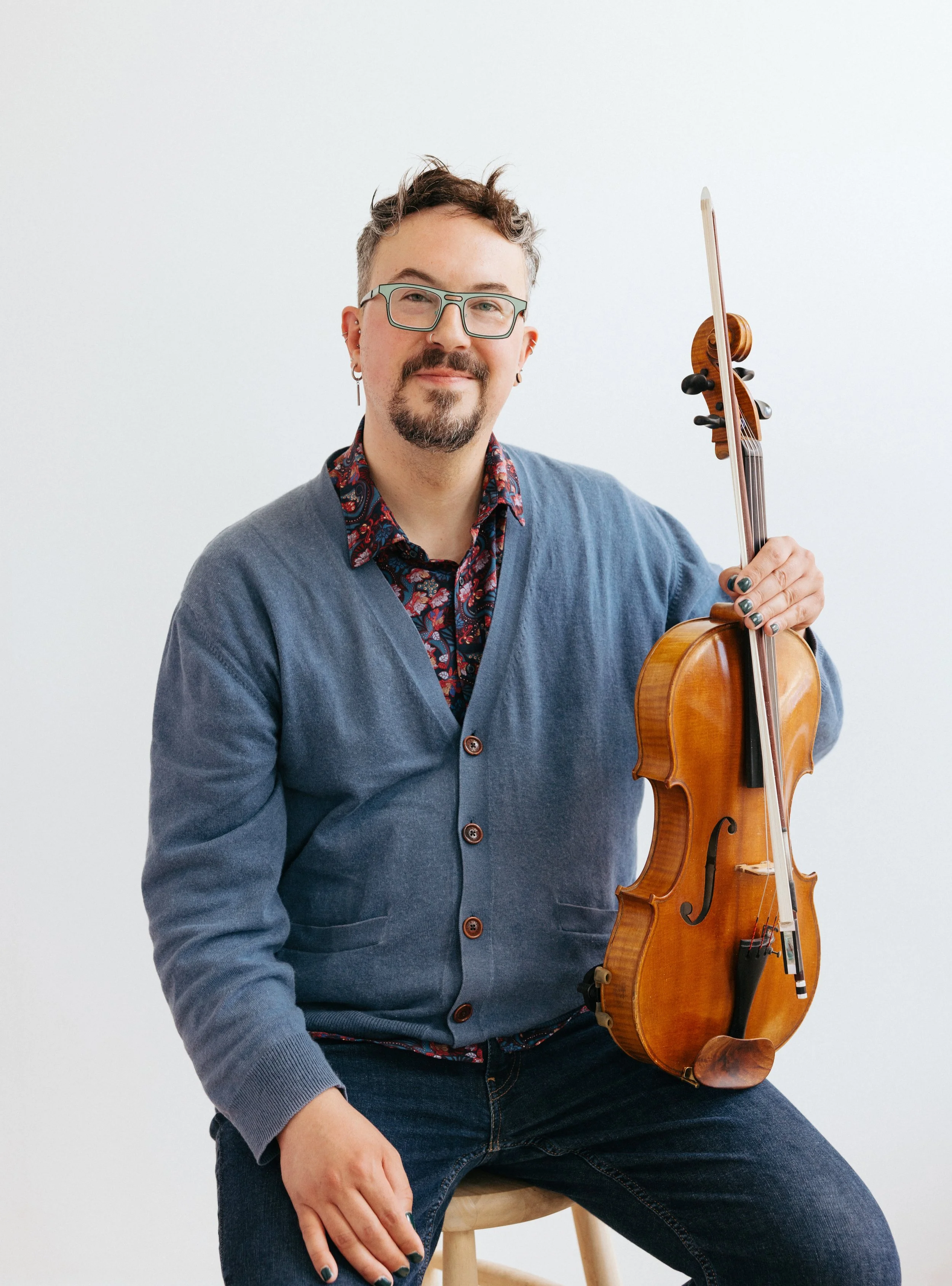 A person with short, curly hair, glasses, and a beard, wearing a blue cardigan over a patterned shirt, sitting on a wooden stool, holding a viola in one hand, against a plain white background.