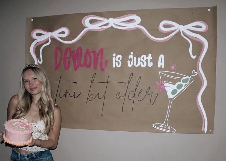 A young girl holding a pink birthday cake with a sign on the wall behind her that reads 'Devon is just a tiny bit older' with decorative pink and white ribbons and a cartoon cocktail glass with green olives.
