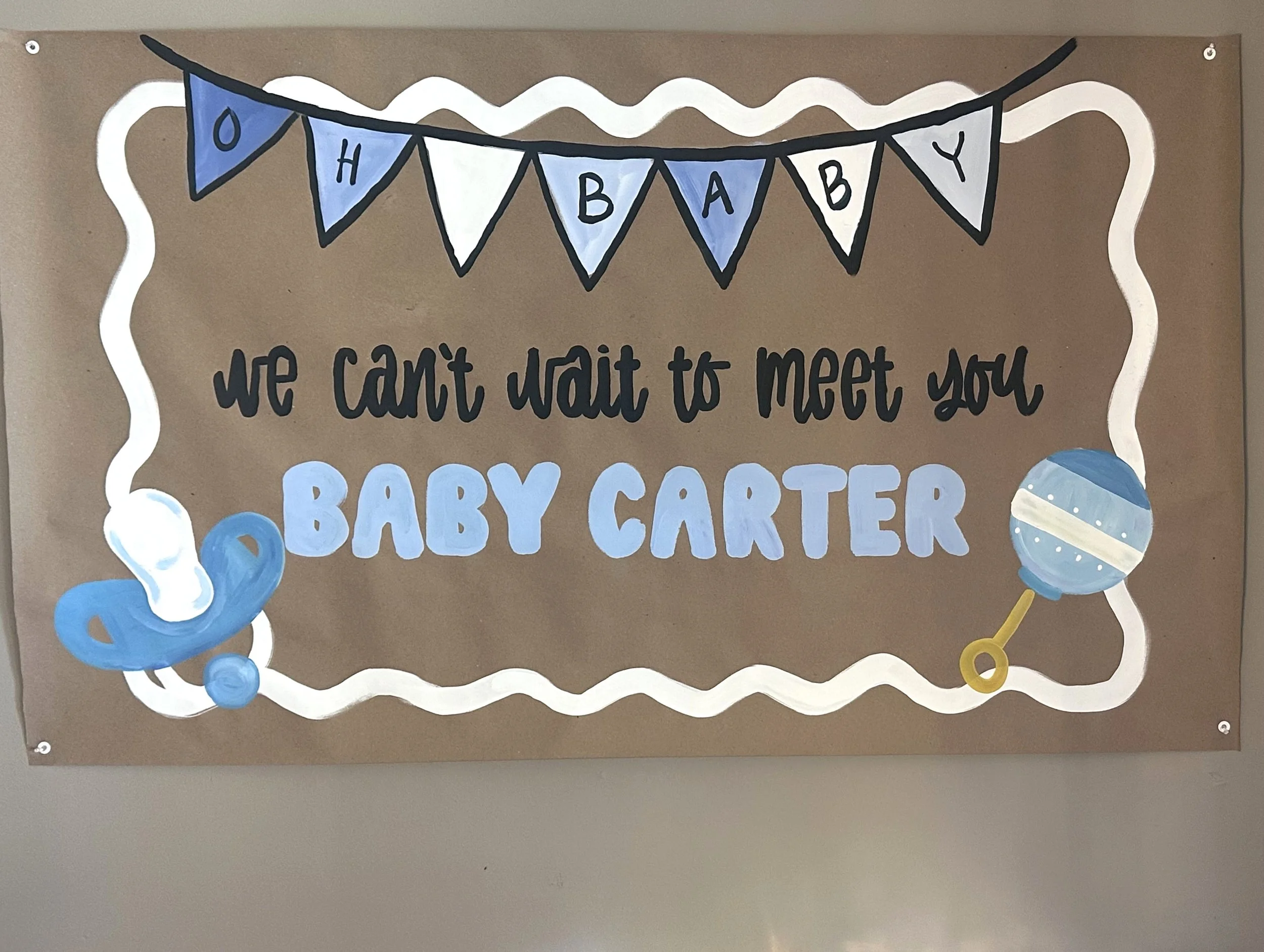 Decorative sign with a blue and white banner that reads 'O H A B A B Y' and handwritten text underneath saying 'we can't wait to meet you Baby Carter.' There are blue and white balloons and a white, wavy border around the sign.
