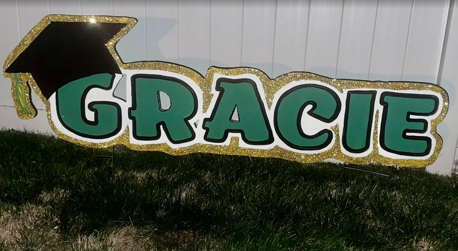Decorative sign with the word 'GRACIE' in large green letters outlined in black, topped with a black graduation cap with a green tassel, bordered with gold glitter, placed on artificial grass.