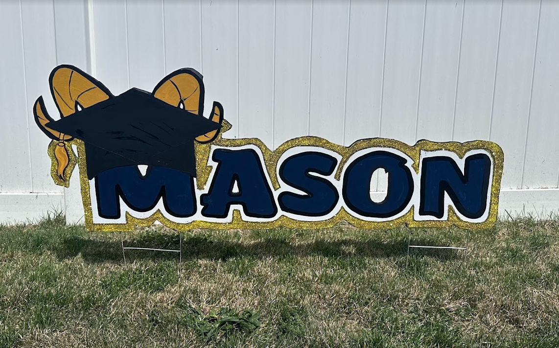 Decorative sign with a cartoon goat wearing a graduation cap and large blue letters spelling 'MASON' on a lawn against a white fence.