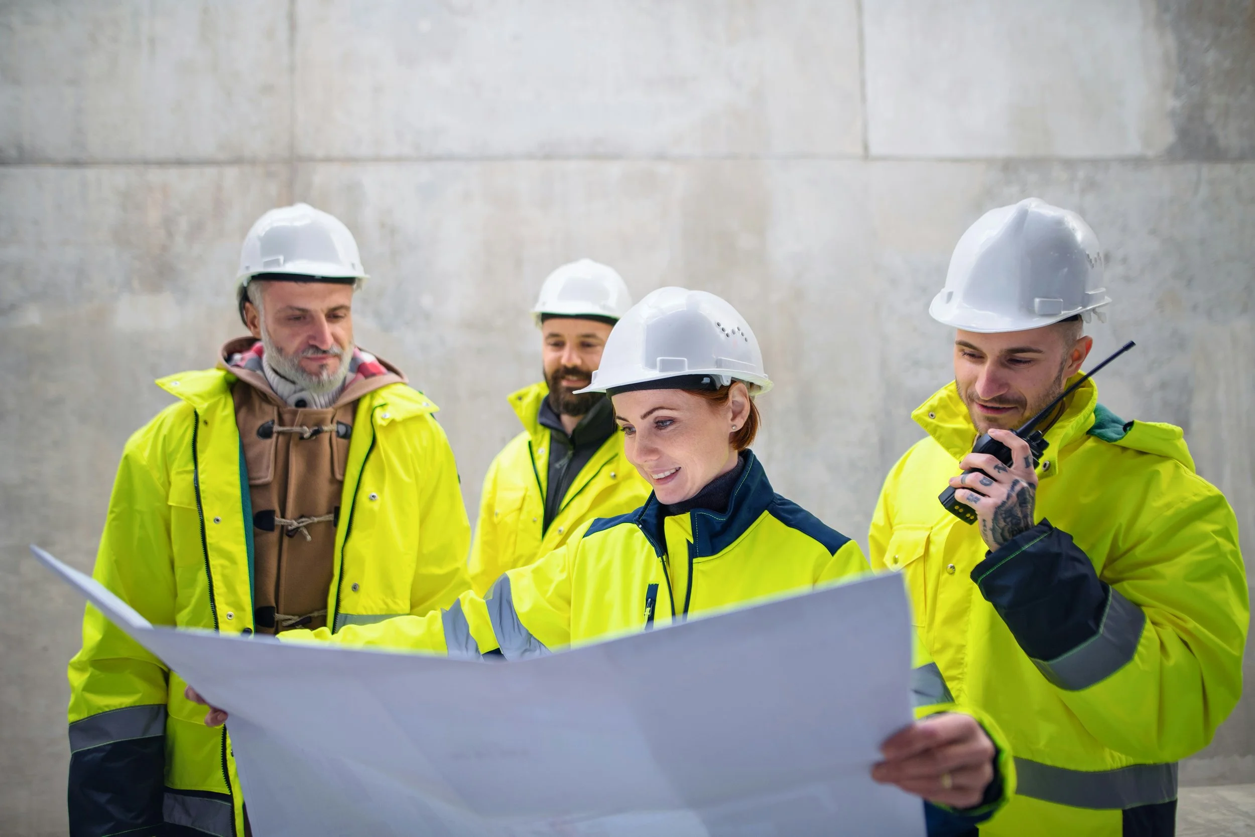 Group of five construction workers wearing yellow safety jackets and white hard hats examining a large blueprint against a concrete wall background.