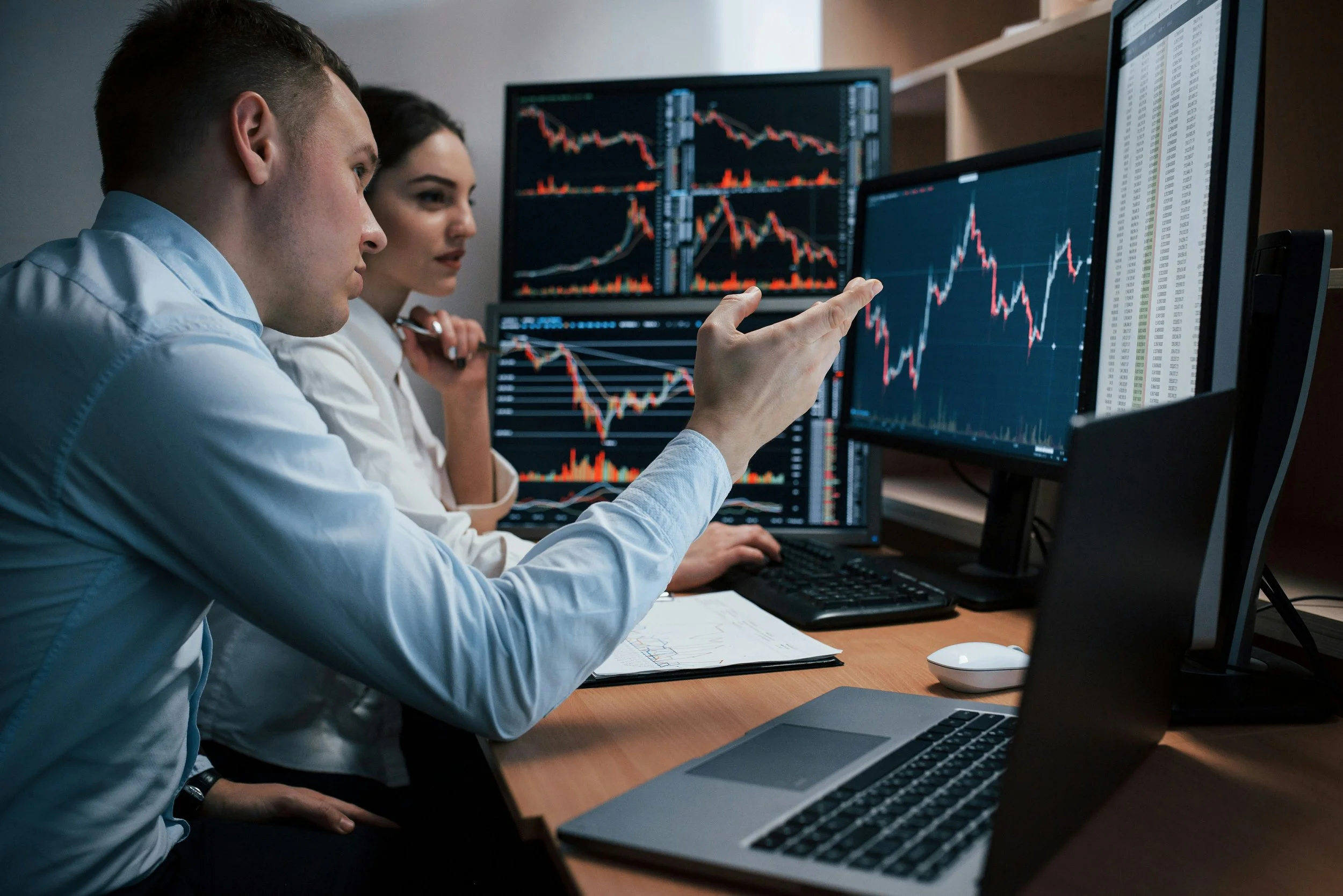 Two individuals analyzing stock market charts on computer monitors in an office setting.