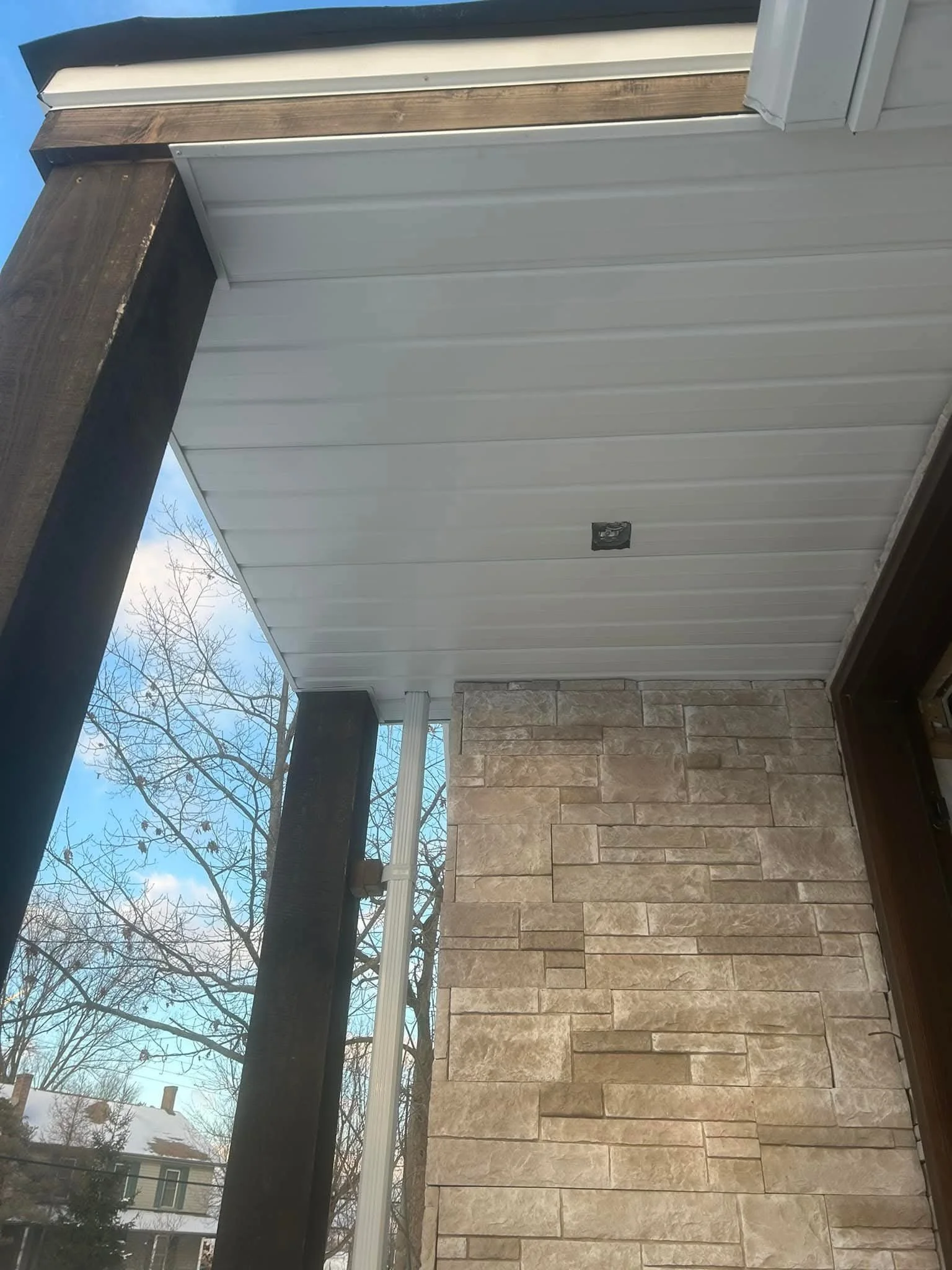 The image shows the exterior side of a building with a stone facade wall, white siding, a window, and a snow-covered yard outside. Part of a black wooden post and gutter pipe are also visible.