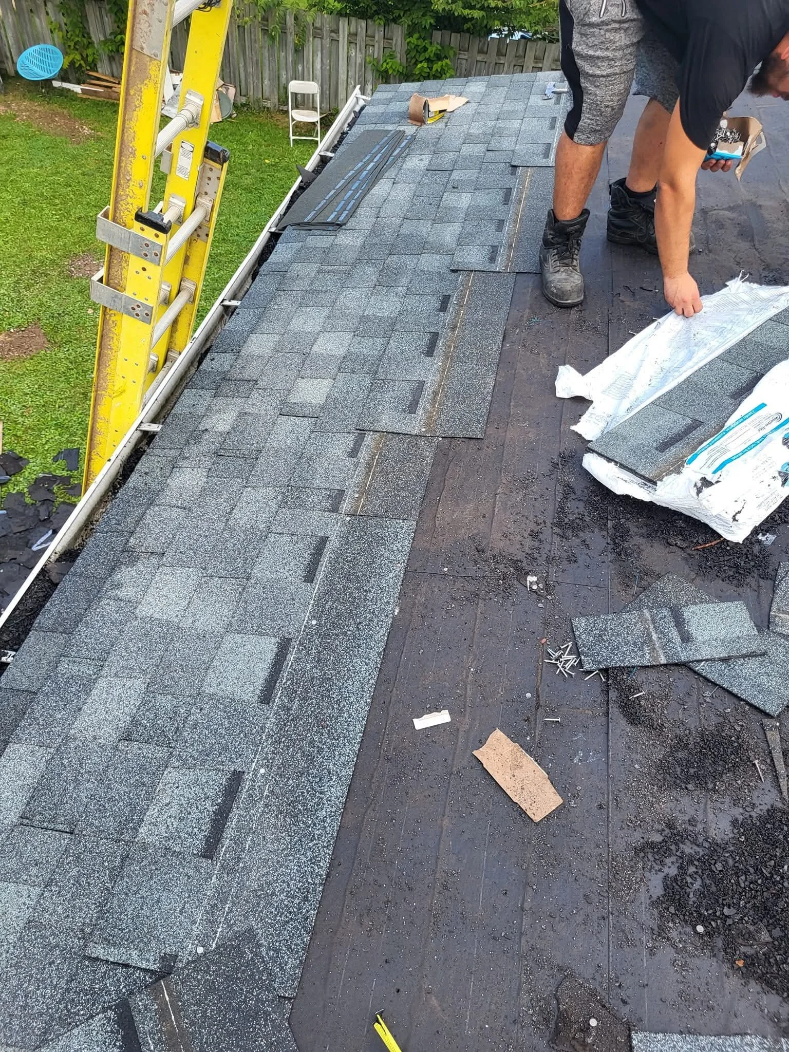 A person installing asphalt shingles on a roof. Ladders and tools are nearby, and the person is wearing work boots, bent over working on the shingles.
