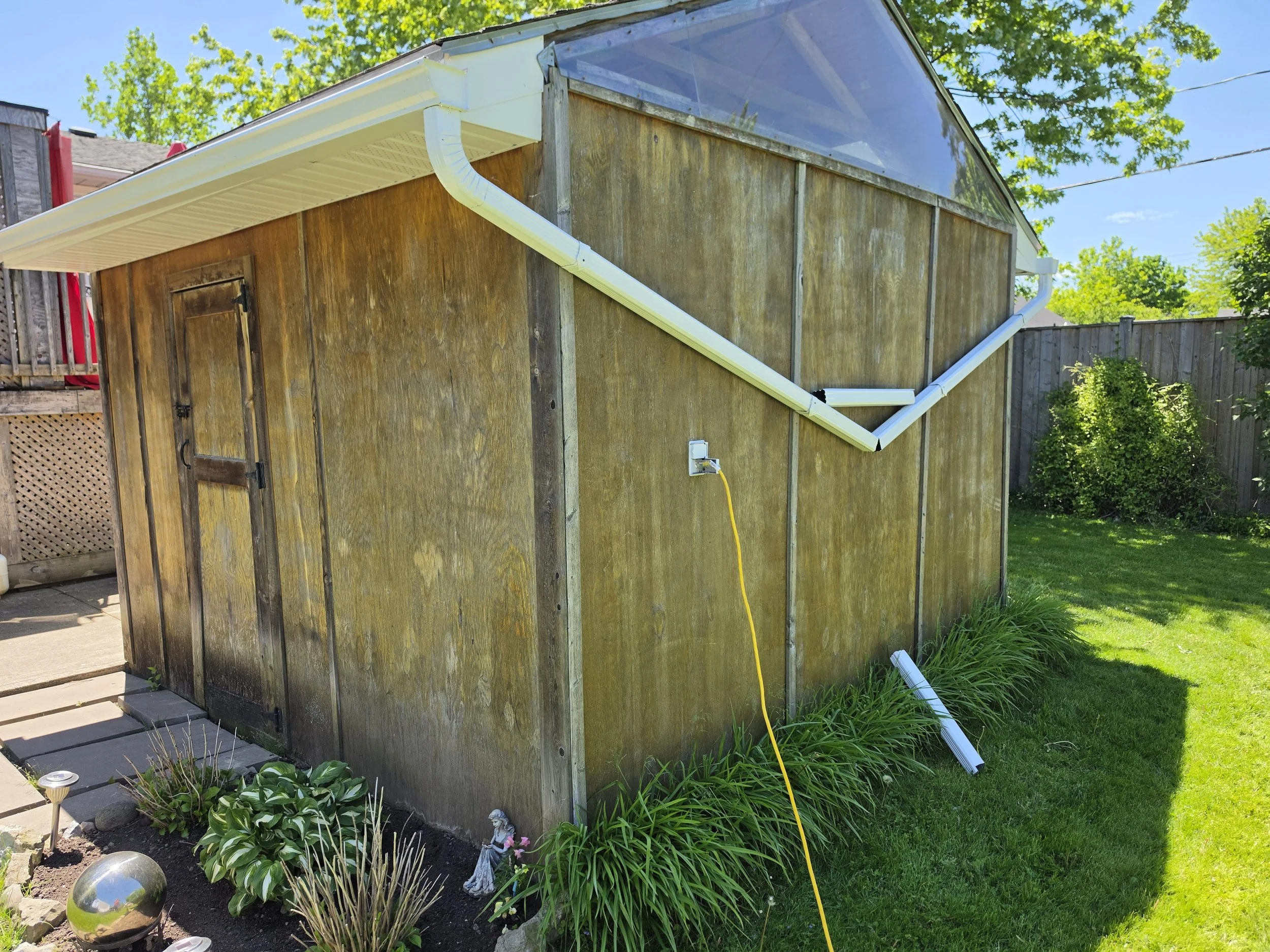 A backyard wooden shed with a new white gutter system installed, including a downspout. The shed is surrounded by green grass, plants, and decorative garden ornaments. The sky is clear and sunny.