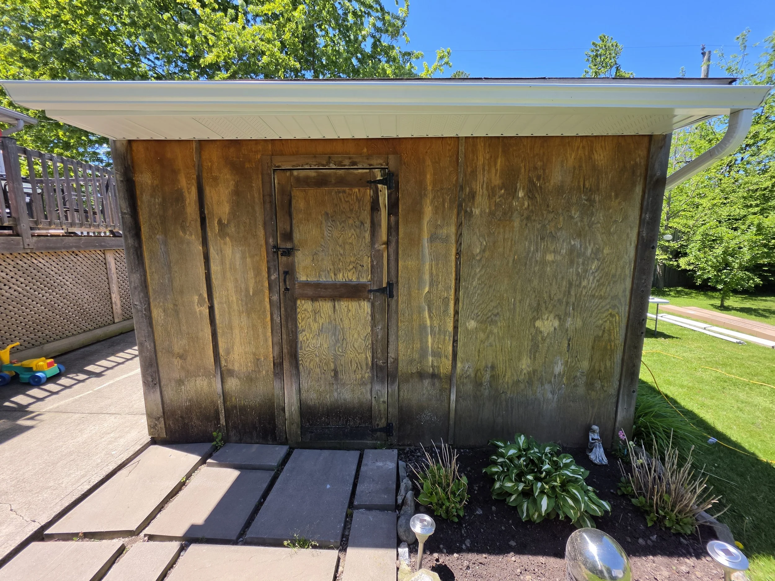Image of the back of a small wooden shed with a closed door, weathered wood panels, and a white metal roof, located in a landscaped yard with plants, garden decor, and a paved walkway, with green trees and a blue sky in the background.