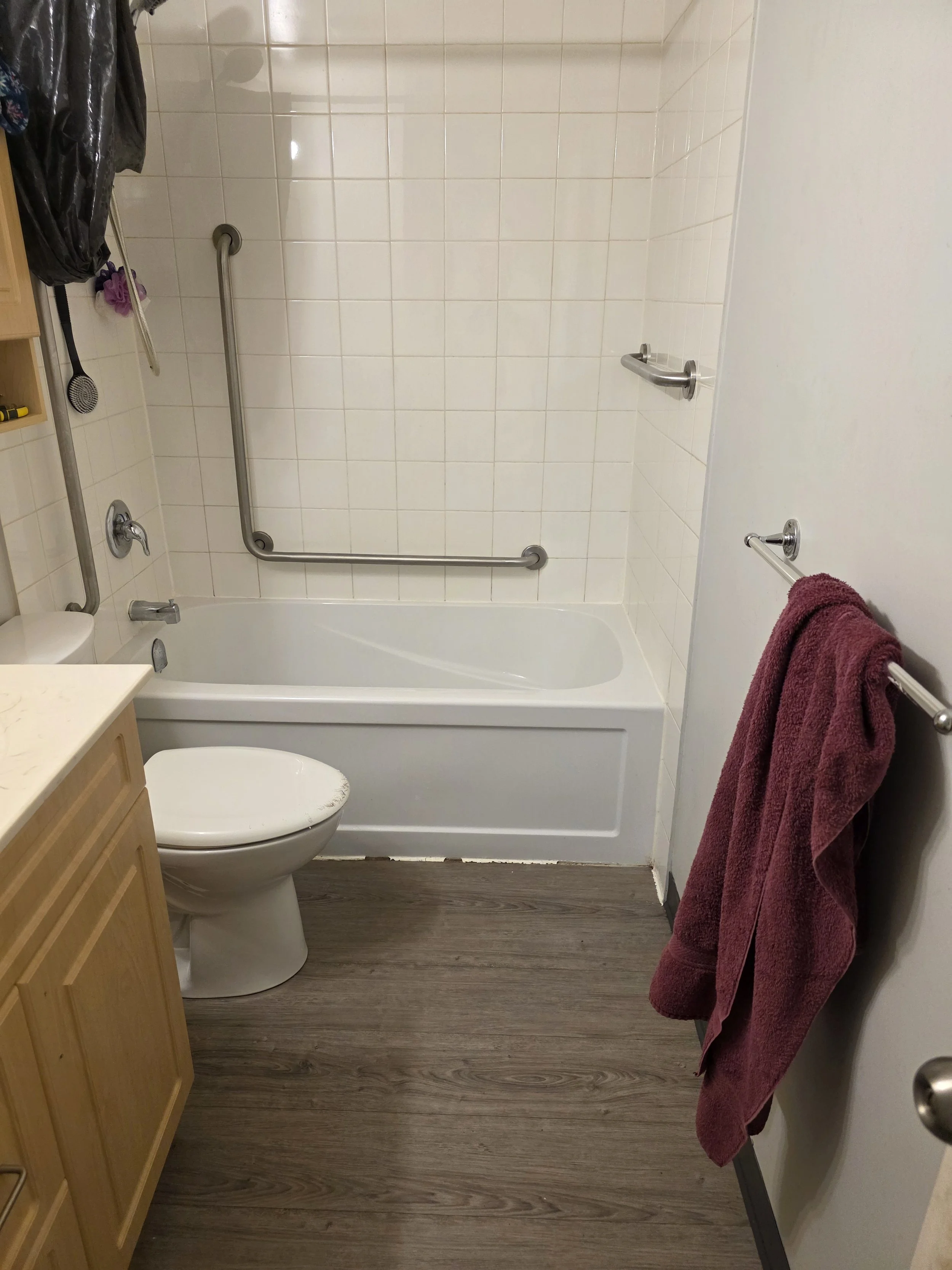 This is a small bathroom with a white bathtub, a toilet, a wooden cabinet, a towel rack with a maroon towel, and plastic grab bars on the wall for safety. The flooring is wood-like and the walls are tiled in white.
