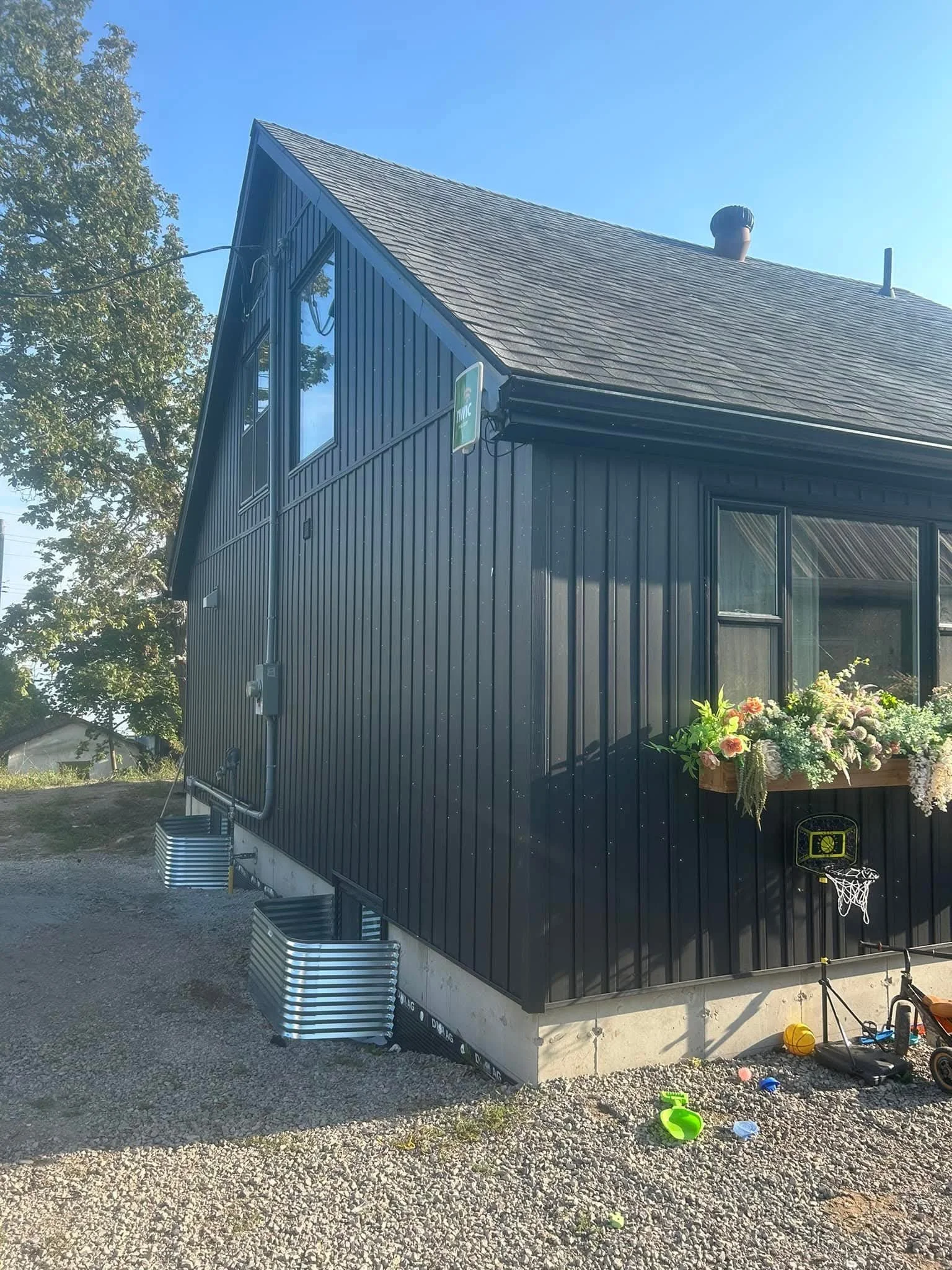 A black modern house with a steep roof and large windows, a flower box with colorful flowers, a small basketball hoop, and building materials and toys on the gravel ground outside.