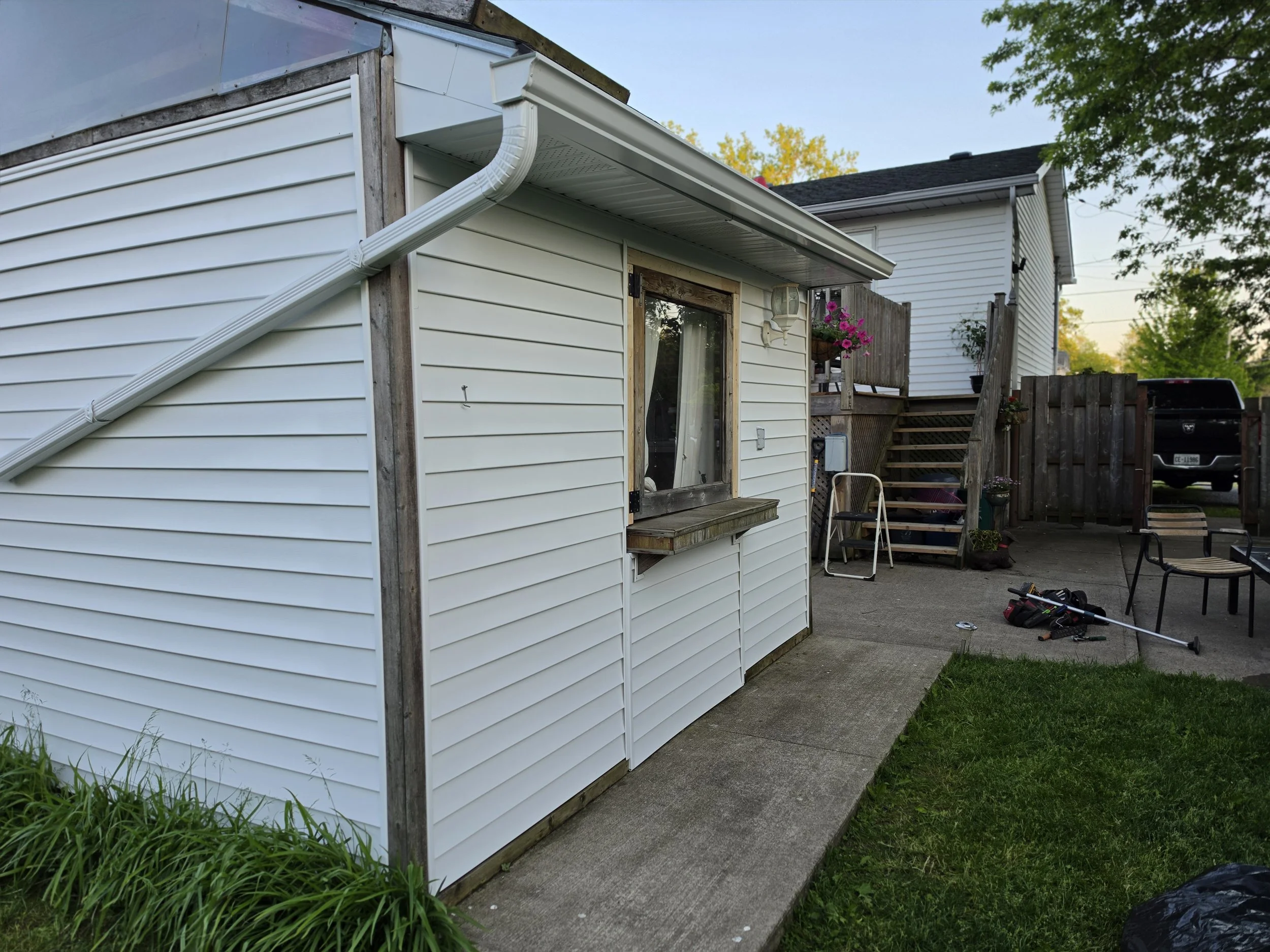 View of a residential backyard with a white house siding, a concrete patio, outdoor chairs, tools on the ground, and a wooden deck with stairs leading up to the house, surrounded by trees and a fence.