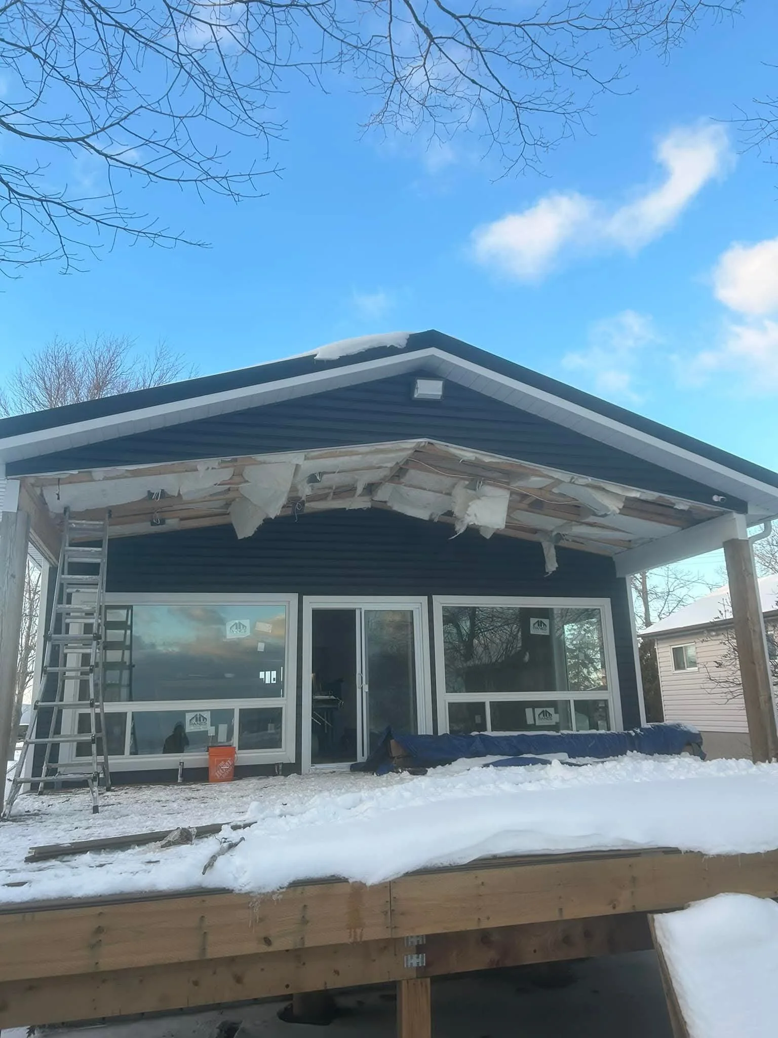 A house under construction with a partially completed roof, wooden framing, and snow on the ground and roof. There is a ladder leaning against the house and construction materials present.