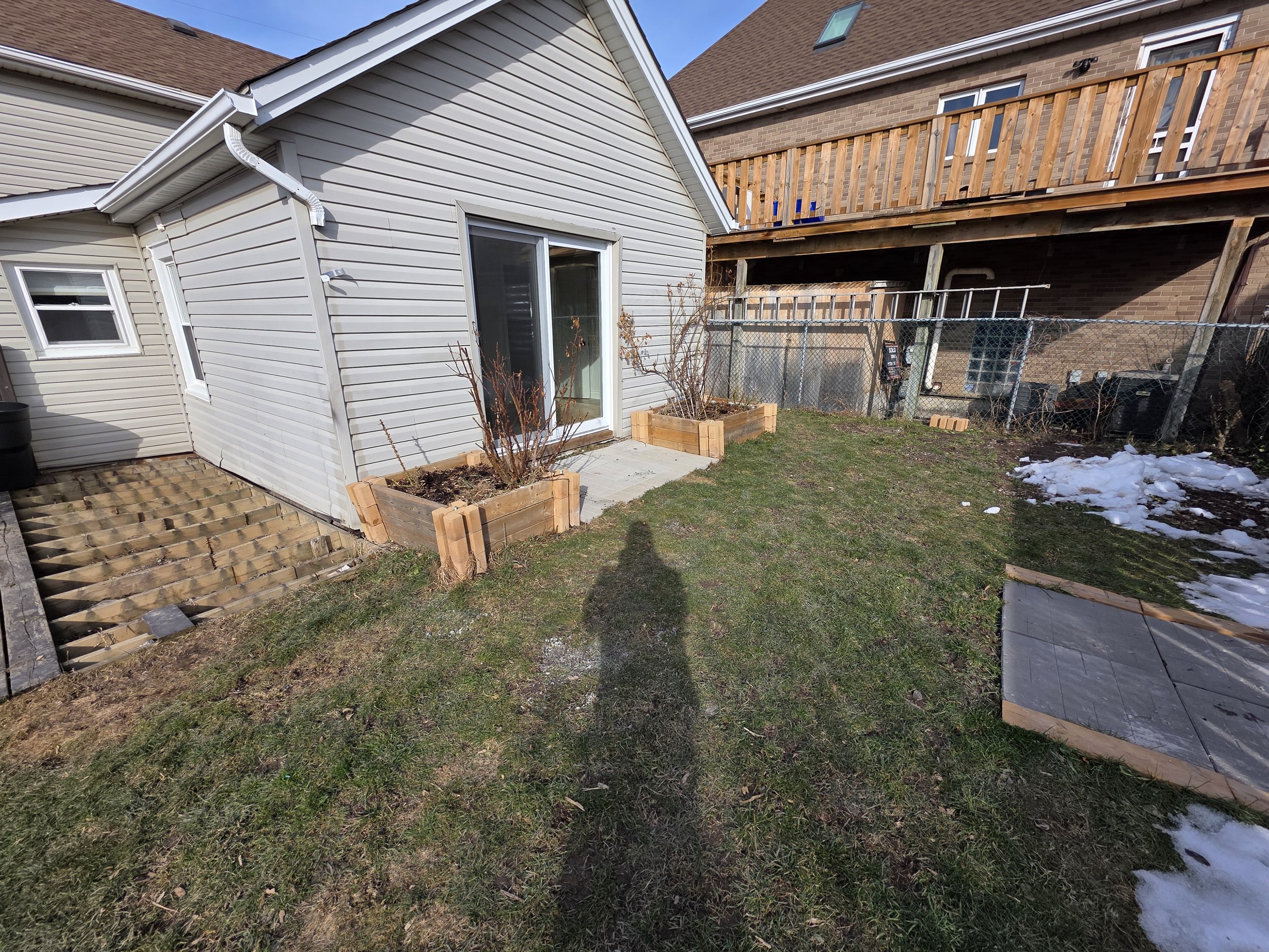 Backyard scene showing a small white house with a sliding glass door, two raised garden beds, a patch of grass, some snow, and a neighboring brick house with a wooden deck.