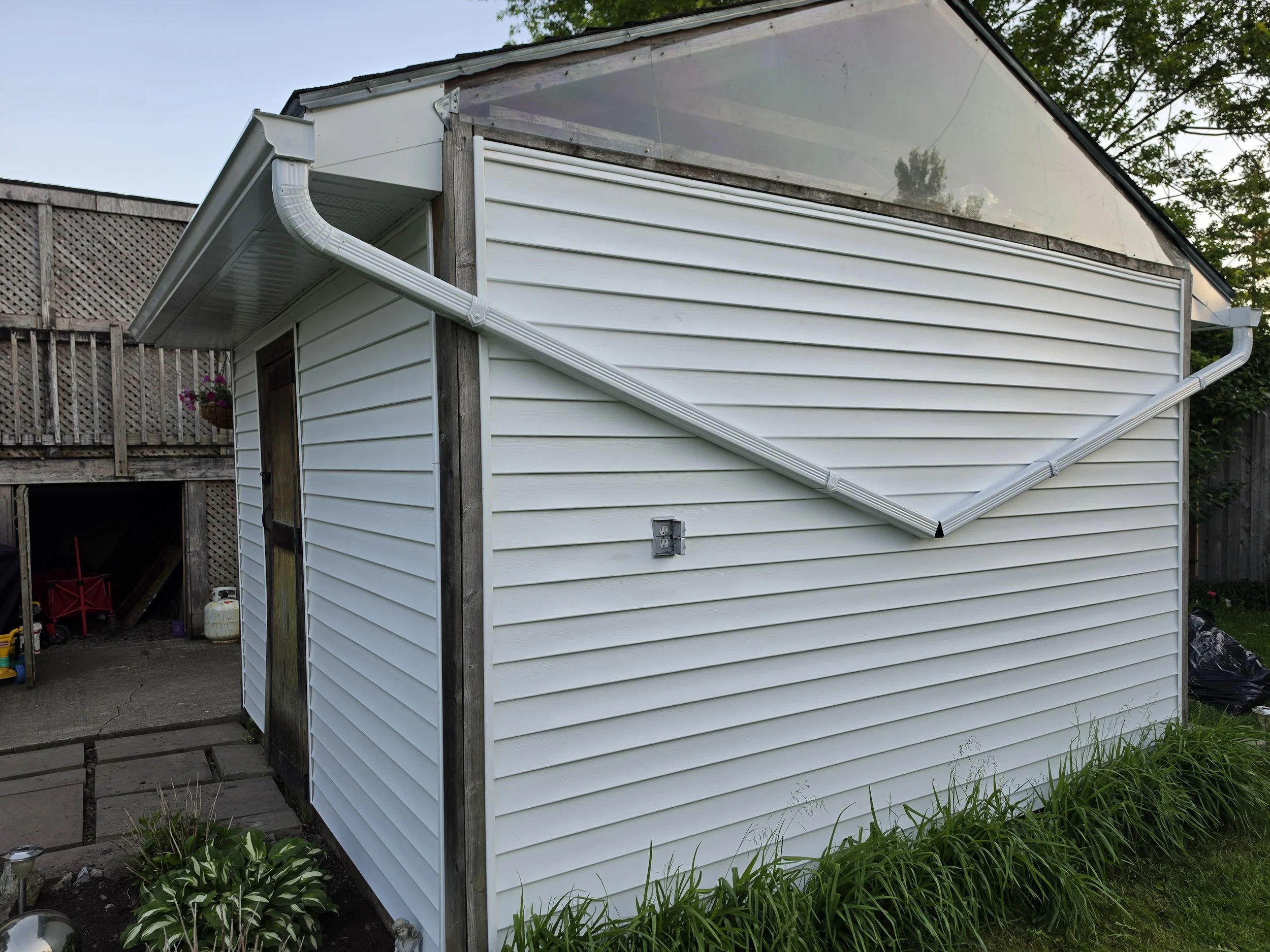 Backyard shed with white vinyl siding, wooden support beams, and a detached roof section with a translucent panel. There are plants and flowers in the surrounding area.