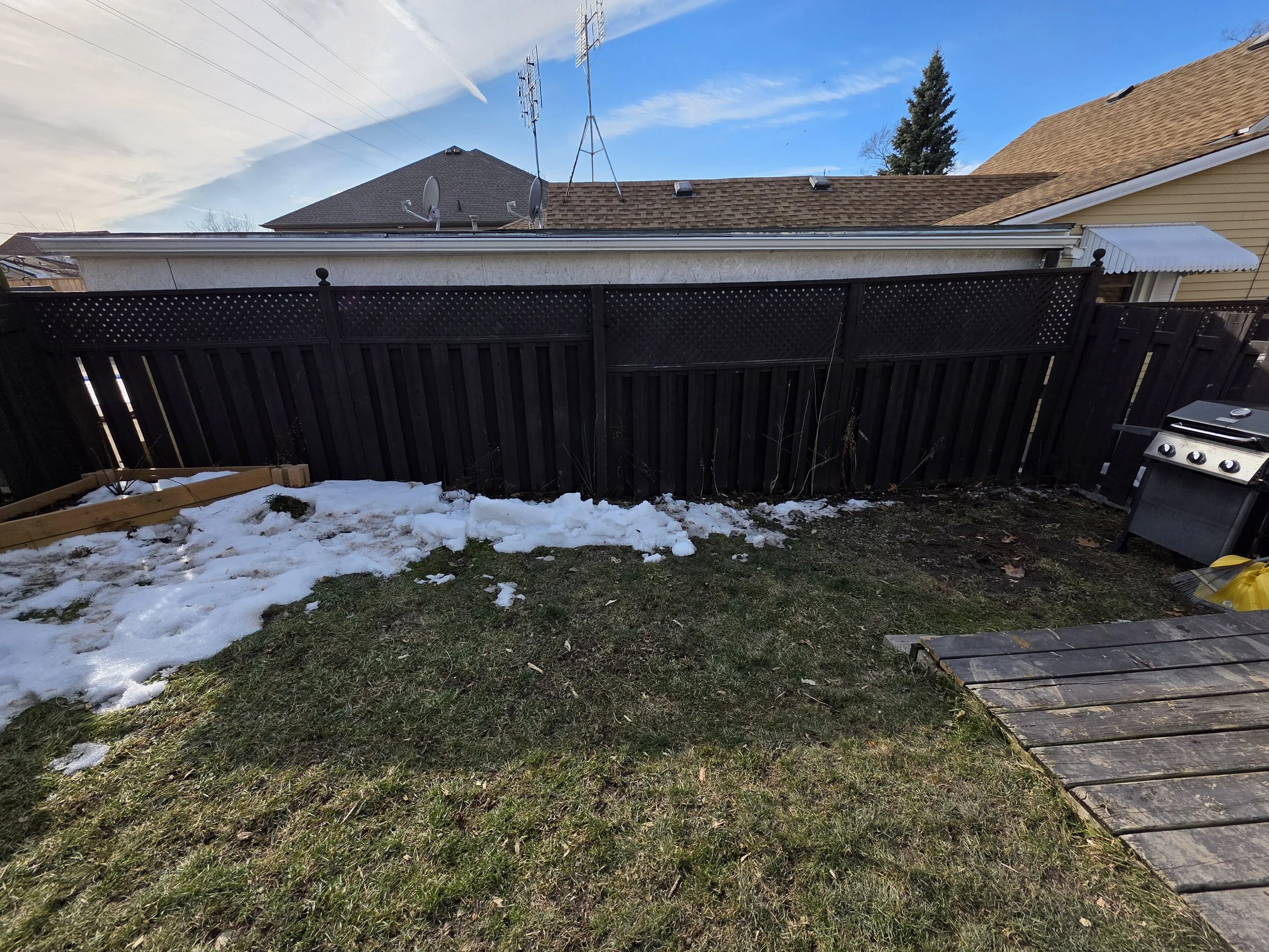 Backyard view with snow patches on grass, a black fence, a barbecue grill in the corner, and neighboring houses with brown and beige roofs under a partly cloudy blue sky.