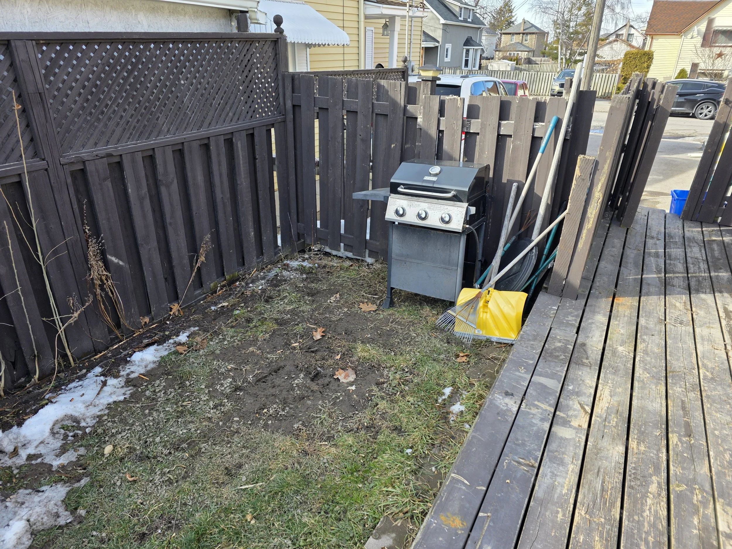 A backyard with a damaged wooden fence, a small barbecue grill, gardening tools, and a yellow snow shovel. The ground has patches of snow and grass, with a city street and residential houses visible in the background.