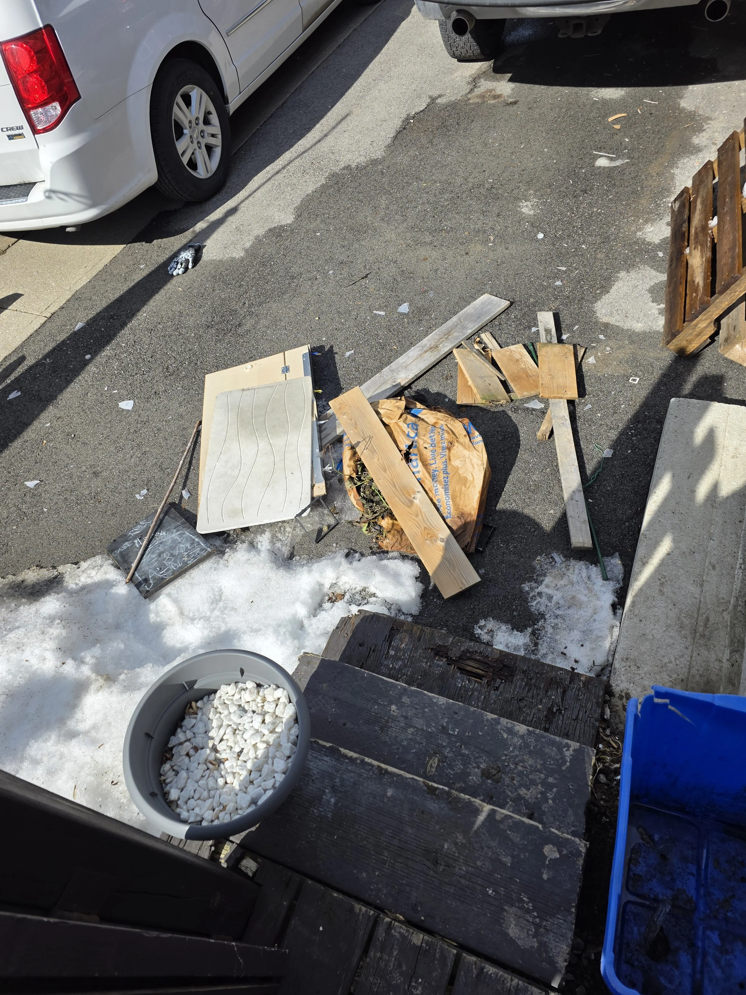 A backyard with a stone patio, a plastic bucket filled with white stones, a wooden planter with soil and dead plants, and part of a deck with weathered wood. Snow is on the ground near the patio, and two cars are parked nearby.