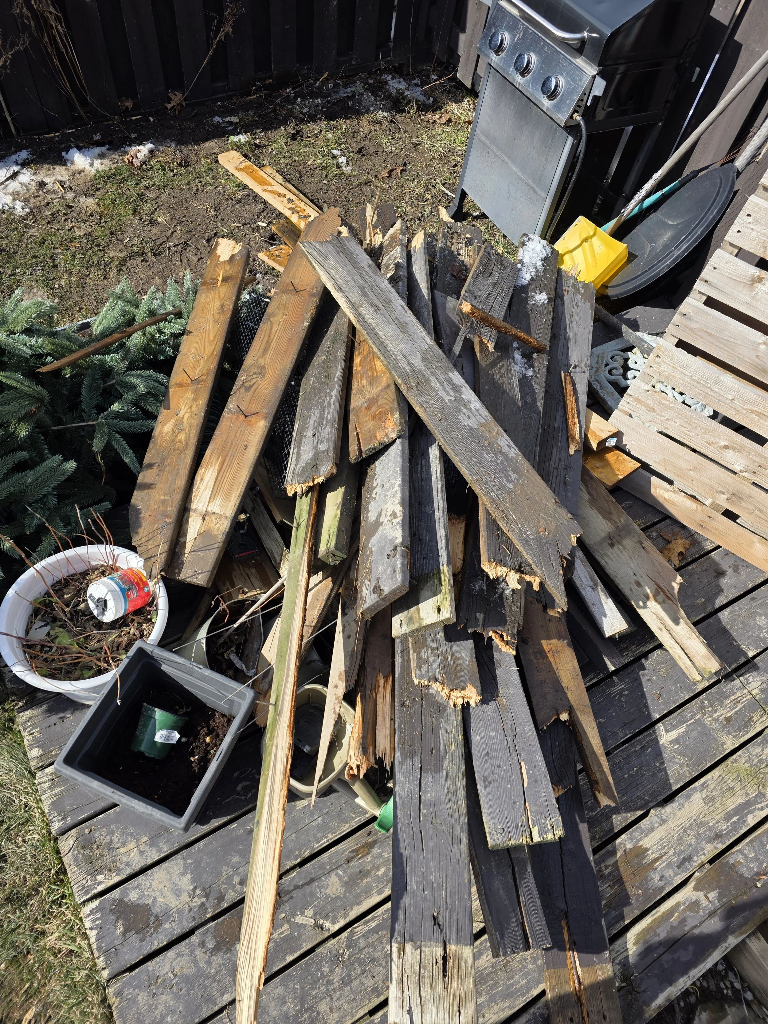 Splintered and broken wooden planks on a wooden deck next to a black grill and gardening tools, with some snow and dirt visible.