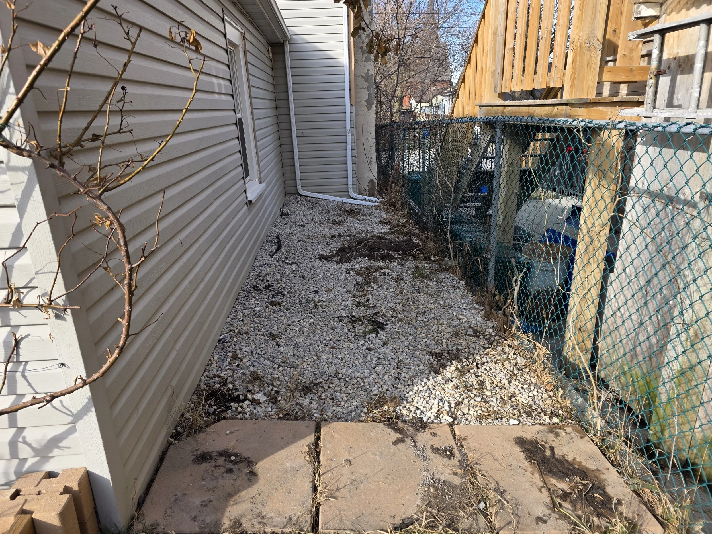 A narrow backyard space between a house with beige vinyl siding and a wooden fence with a gate. The ground is a mix of gravel and dirt with some small plants and debris, and there are a few leafless plants and trees visible in the background.