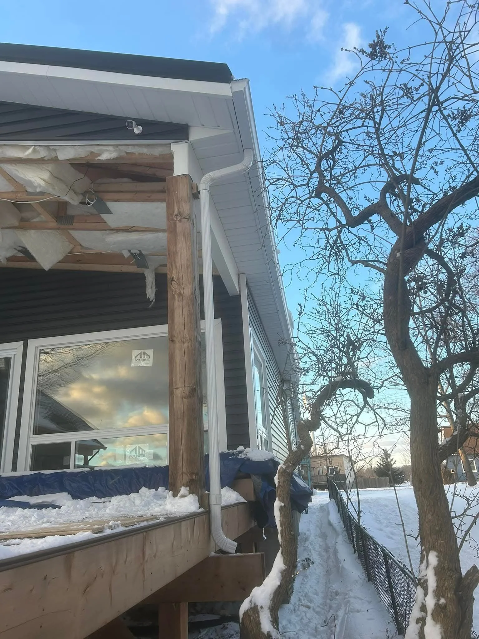 Side view of a house with gray siding and white trim, under construction or renovation, with exposed insulation and building materials. Snow on the ground and on trees, with a cloudy sky above.