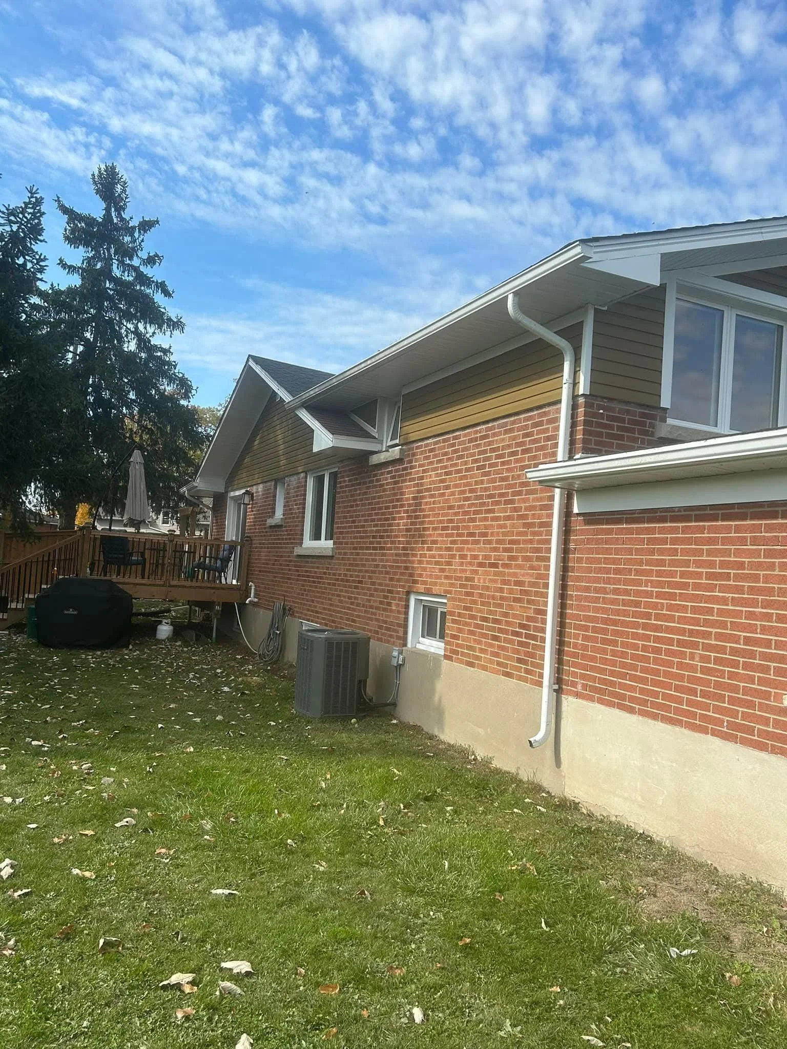 Backyard view showing a brick house with beige siding, a wooden deck with chairs and an umbrella, and a grassy lawn with fallen leaves, under a partly cloudy sky.